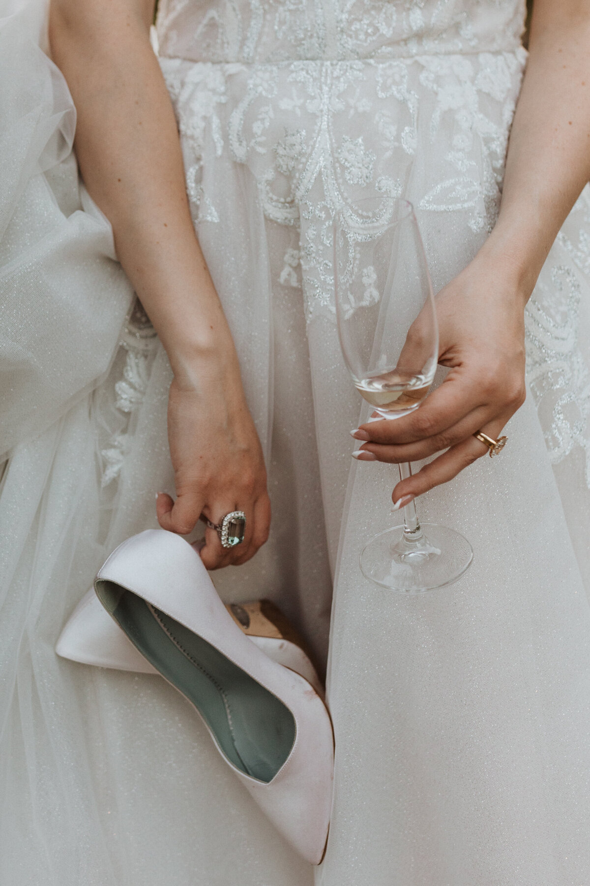 Close up of bride's hands holding champaign glass and white heels in front of wedding dress
