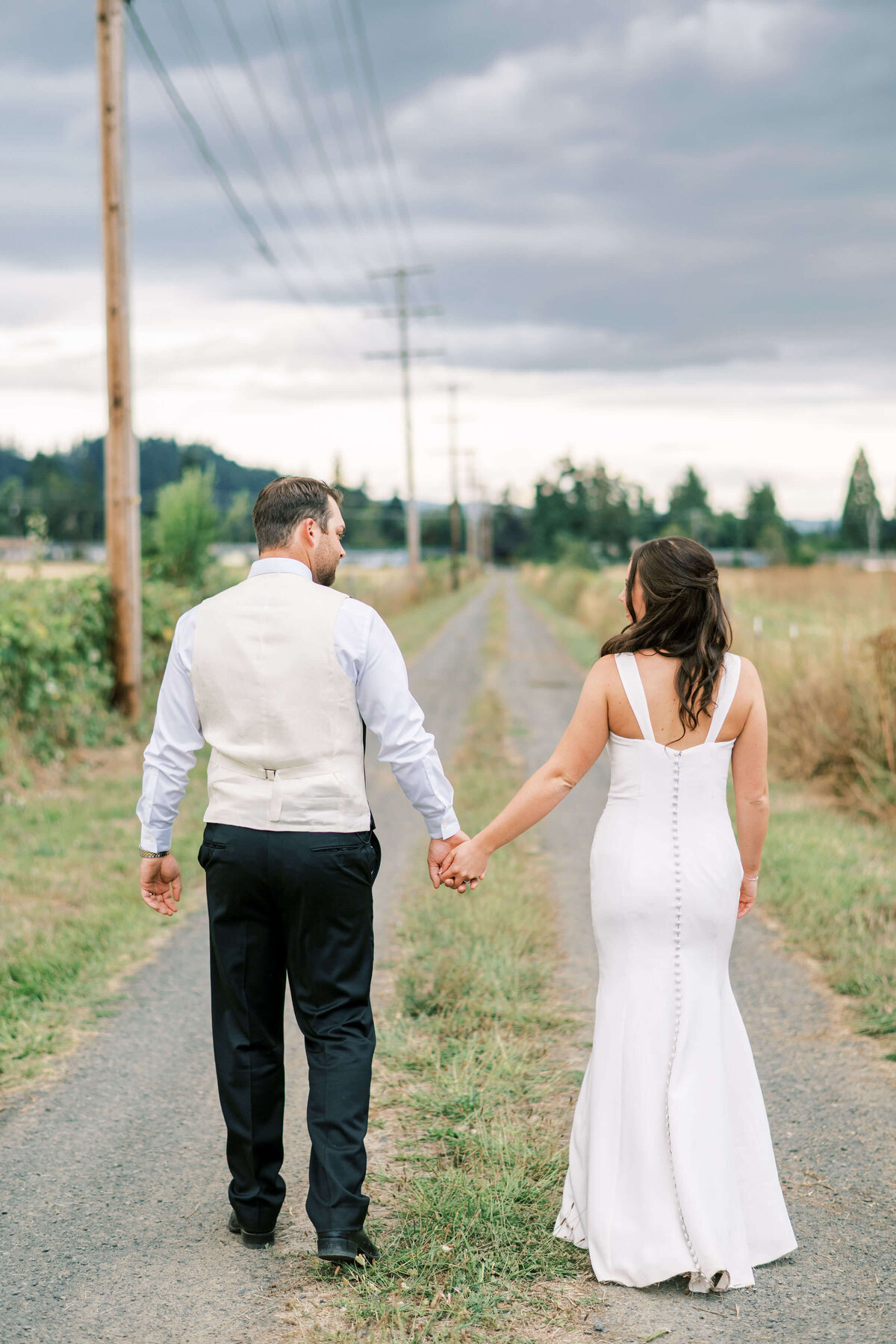 A bride and groom stand outdoors and clasp hands while facing away from the camera.