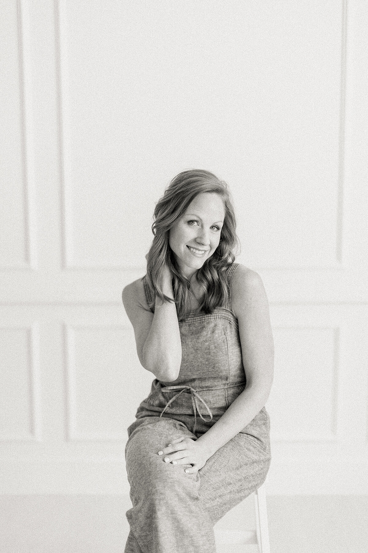 Black and white photo of a Dallas business woman sitting on a stool in a photography studio as she has her hand up behind her neck as she looks and smiles at the camera.