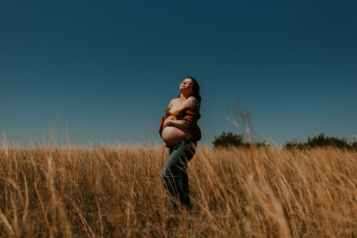 A bare belly pregnant woman standing in a field of grass holding her belly and looking up to the sky in fort worth, texas