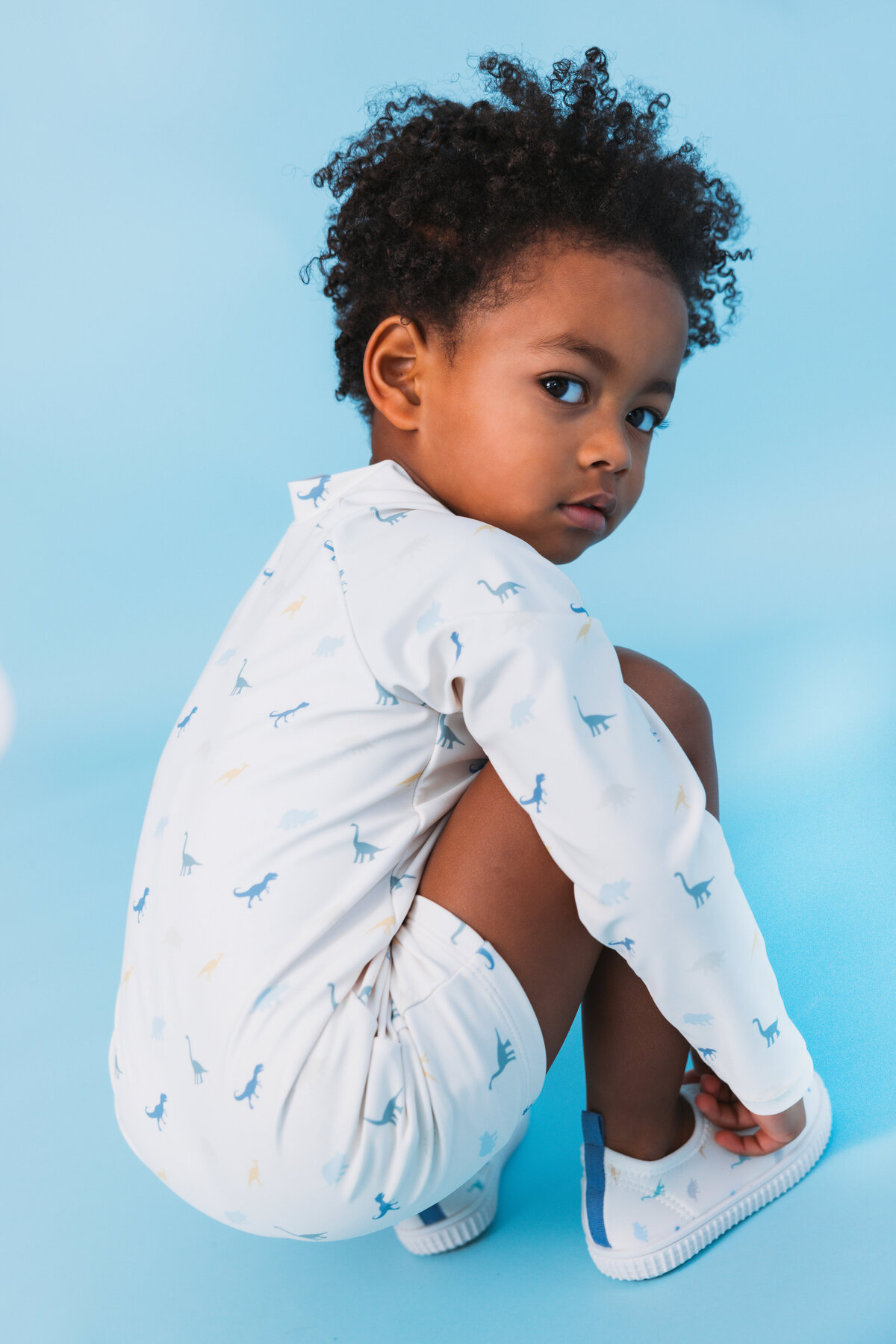 Young child in a white Swim Essentials UV suit with dinosaur print, squatting playfully against a light blue background.