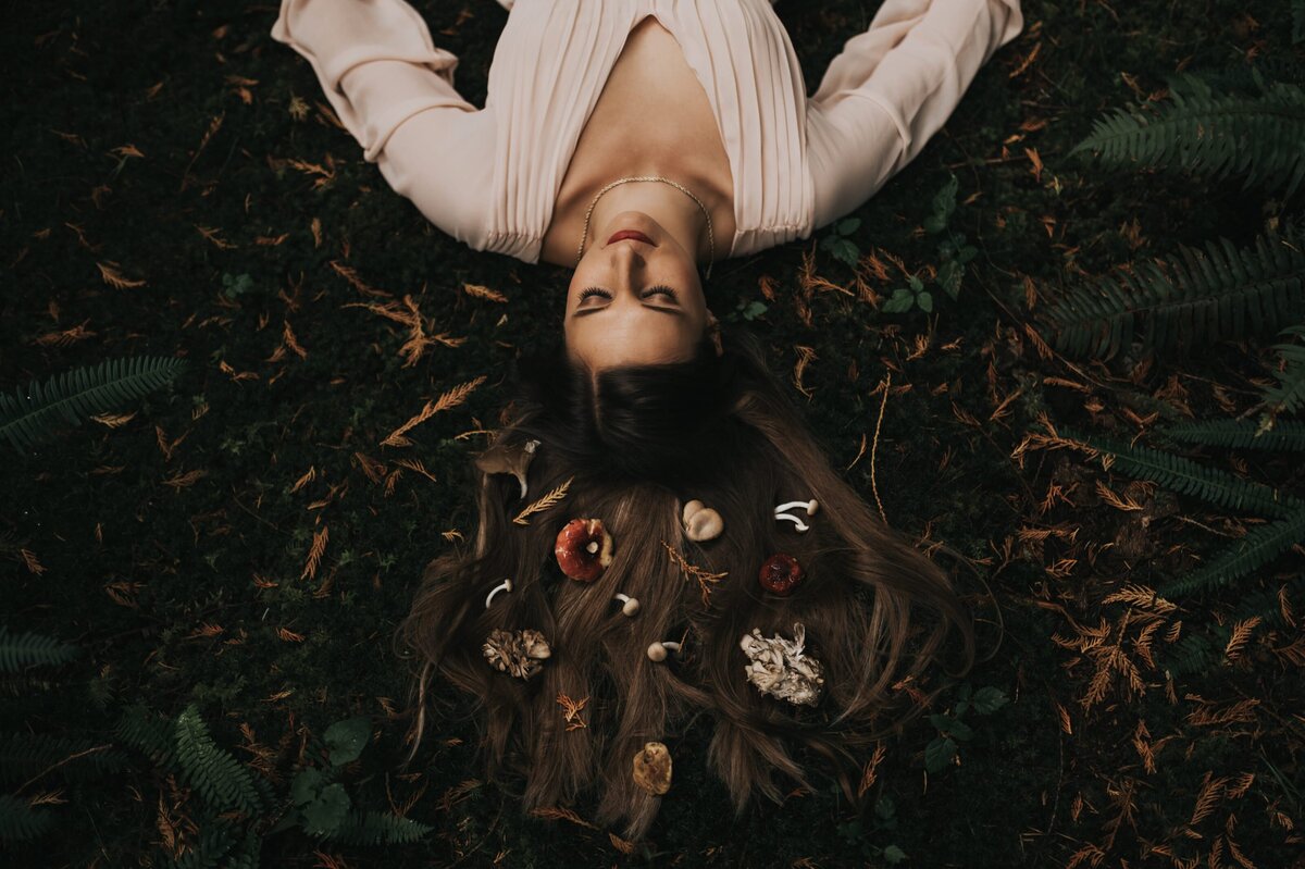 Woman with long, brown hair lays on forest floor with her hands lay by her side. Small mushrooms and foliage are laid flat across her outspread hair.