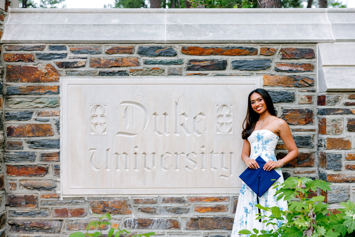 Duke University graduate in blue floral dress holding cap and smiling in front of the Duke sign.