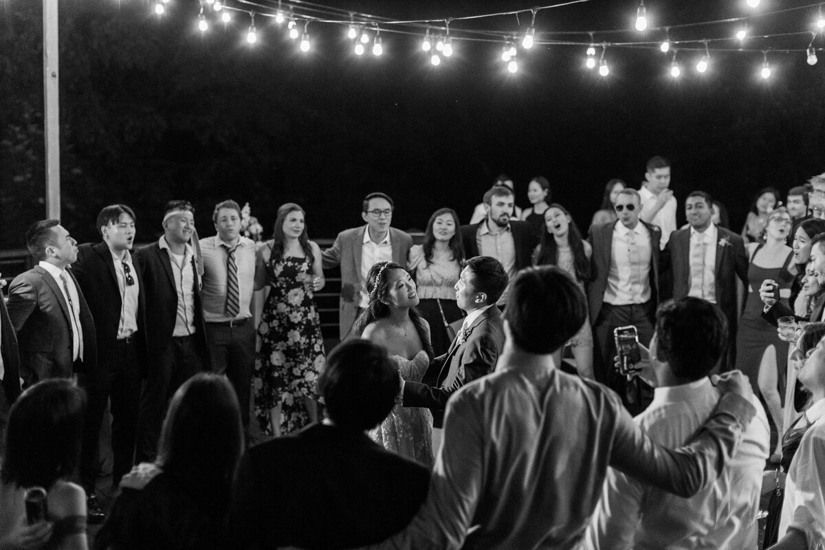 Black and white photo of wedding guests forming a circle around the couple during a group sing-along under string lights at Castle Ladyhawke.