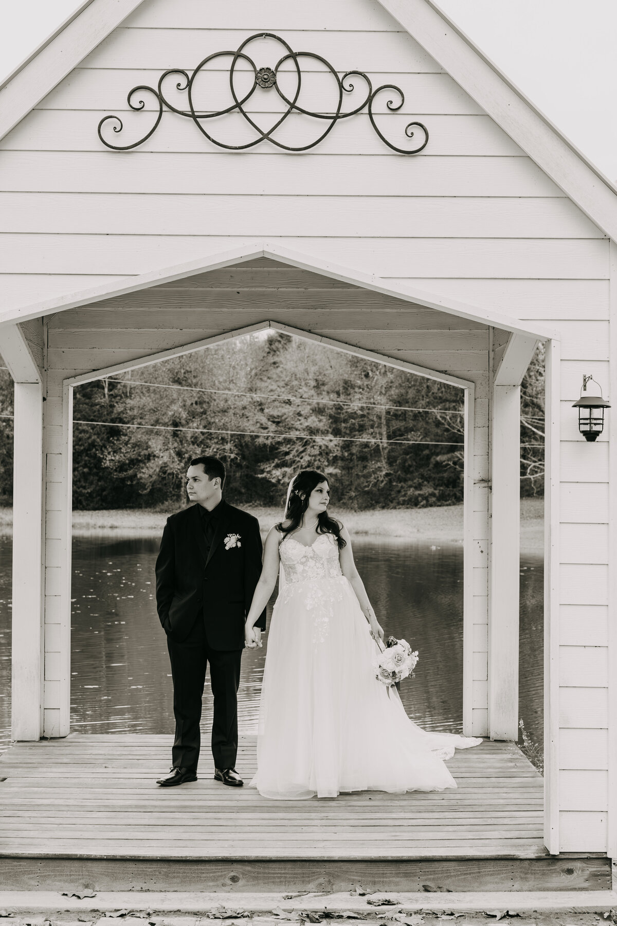 Black and white shot of bride and groom standing under a gazebo on a pond, holding hands, looking away.
