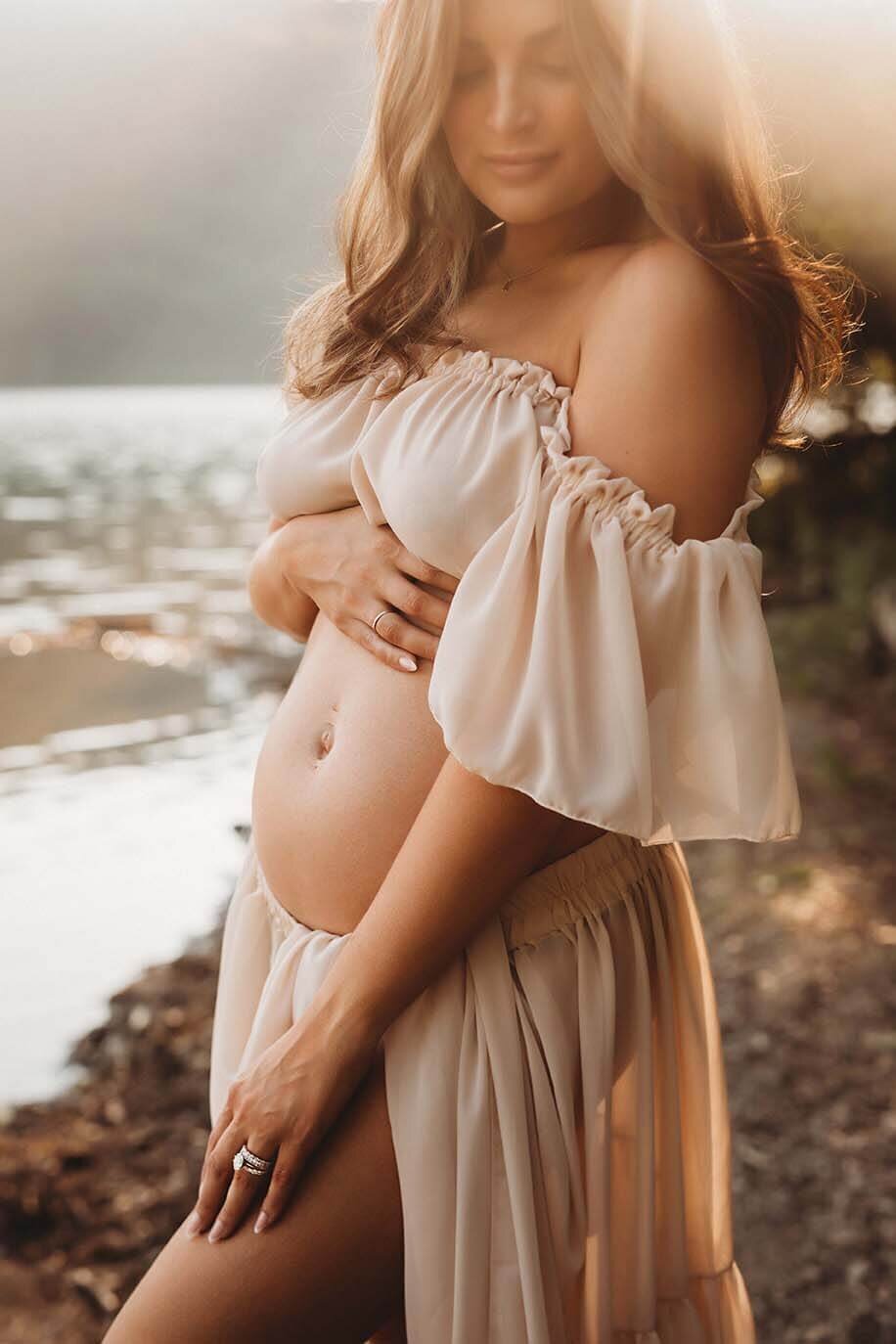A pregnant mother holds her belly while standing on the shore at the lake