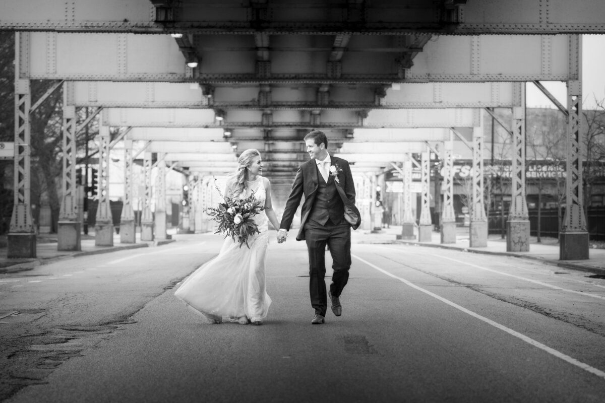 A bride holding her flower and her groom walk hand in hand under a Chicago bridge.
