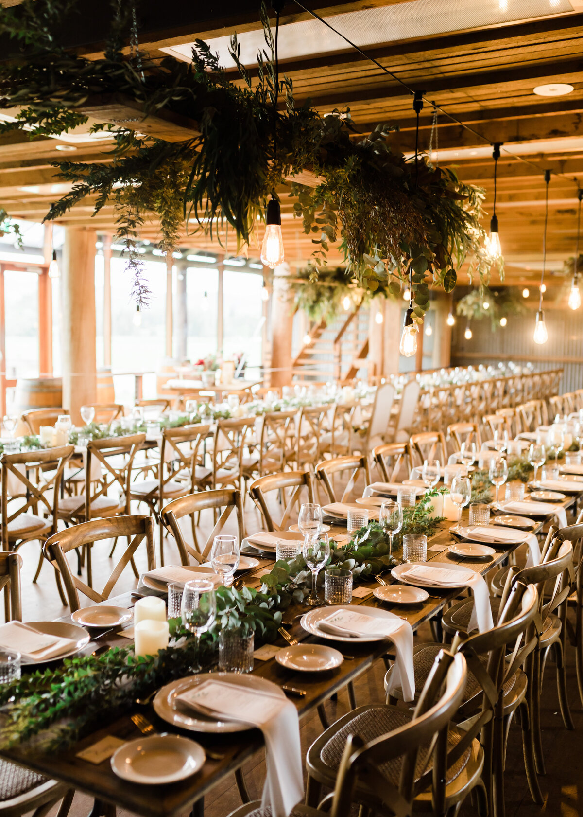 A wedding reception is set up on long wooden tables with a suspended cloud of greenery.