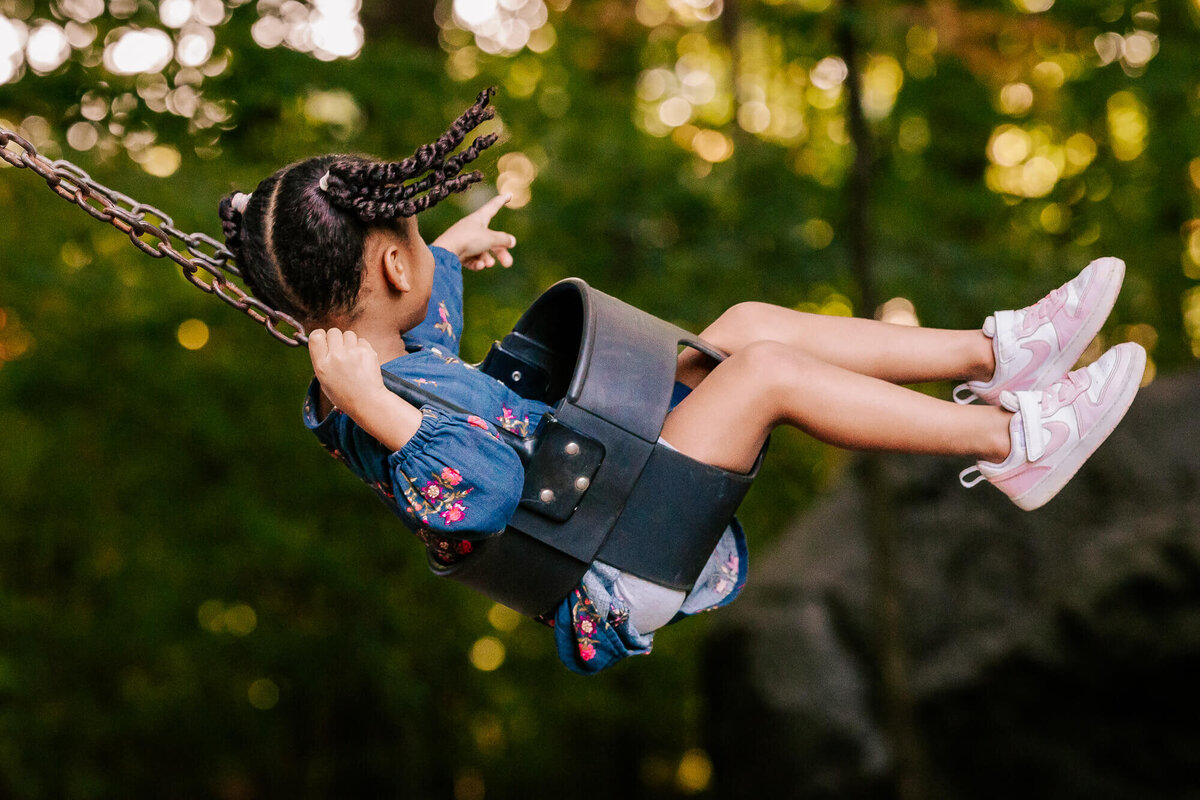 Girl in a blue flowered dress with two black braids swings high into the air, green forest behind her. She's pointing into the woods and we can't see her face.