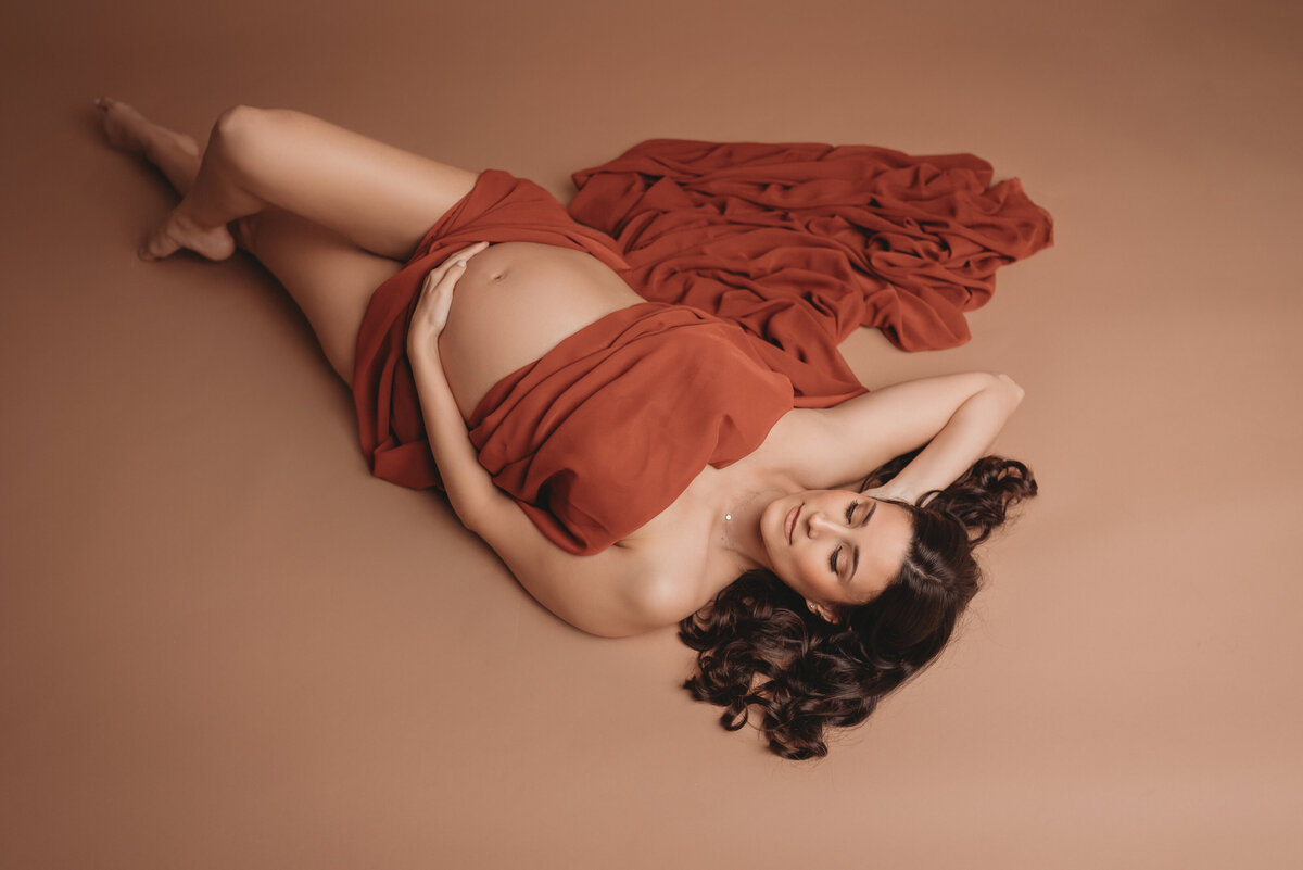 Pregnant woman laying on back draped in burnt orange chiffon fabric legs crossed with one hand on stomach and other hand behind her head