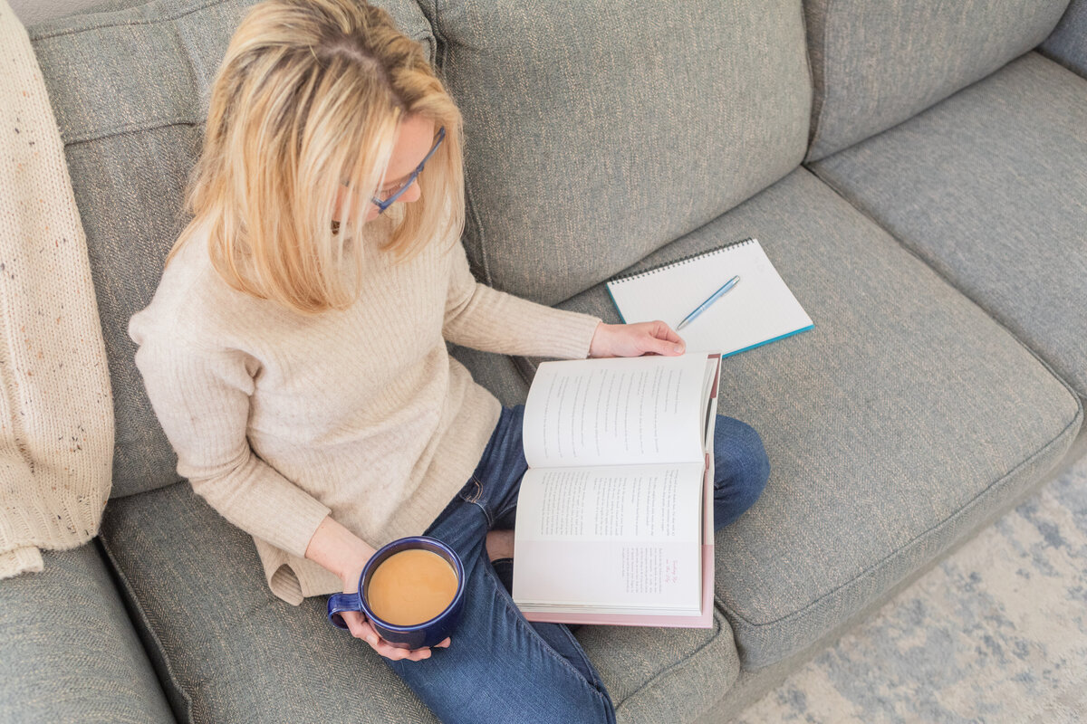 A relaxed business owner sits cross-legged on a couch, reading a book with a blue mug in hand and a notebook nearby—ideal for coaches and entrepreneurs creating content from home.