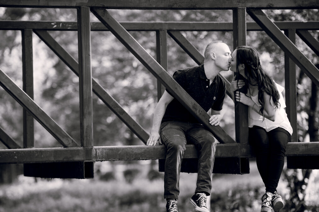Black and white photo of a couple kissing on a bridge.