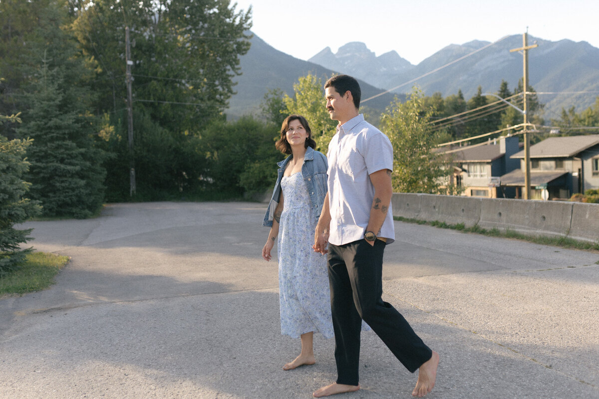 couple walk on tree lined street in Fernie with mountains rising in the bckground