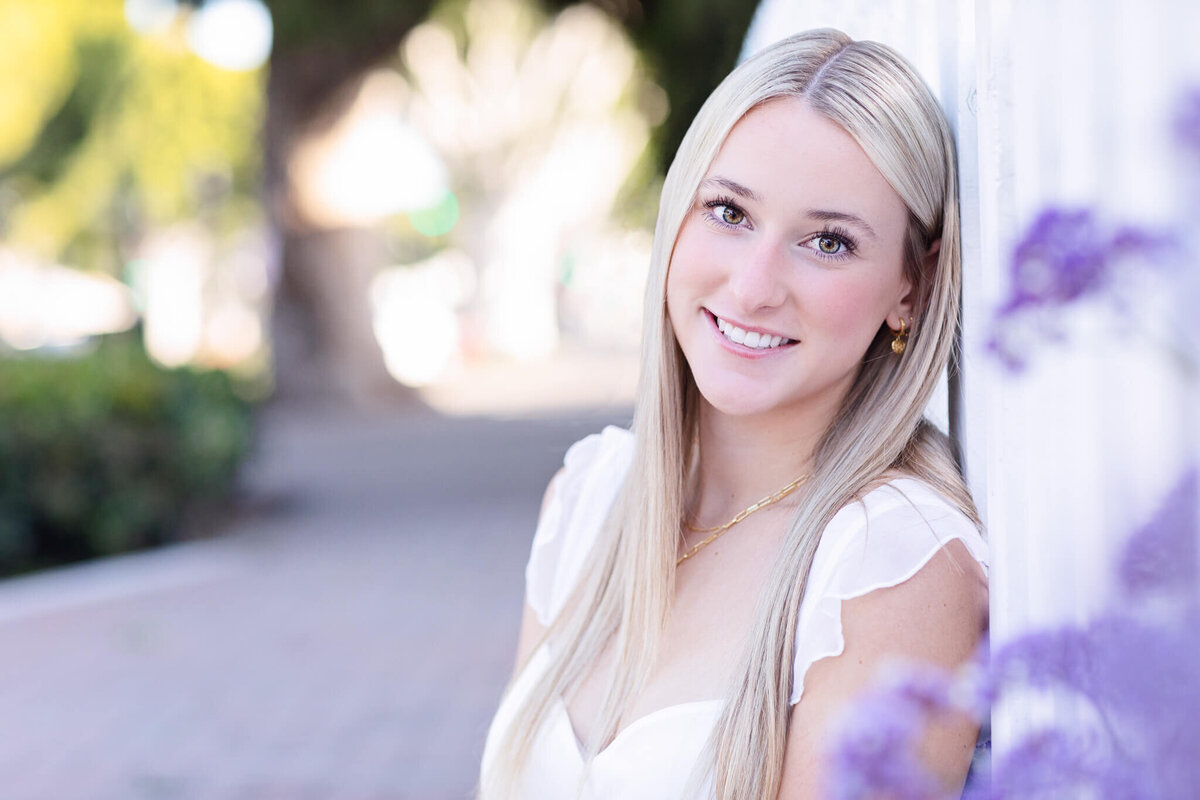 A close-up of a young woman with long, blonde hair sitting on a sidewalk and leaning against a white fence, smiling at the camera with her head leaning against it with purple flowers in front of her out of focus.