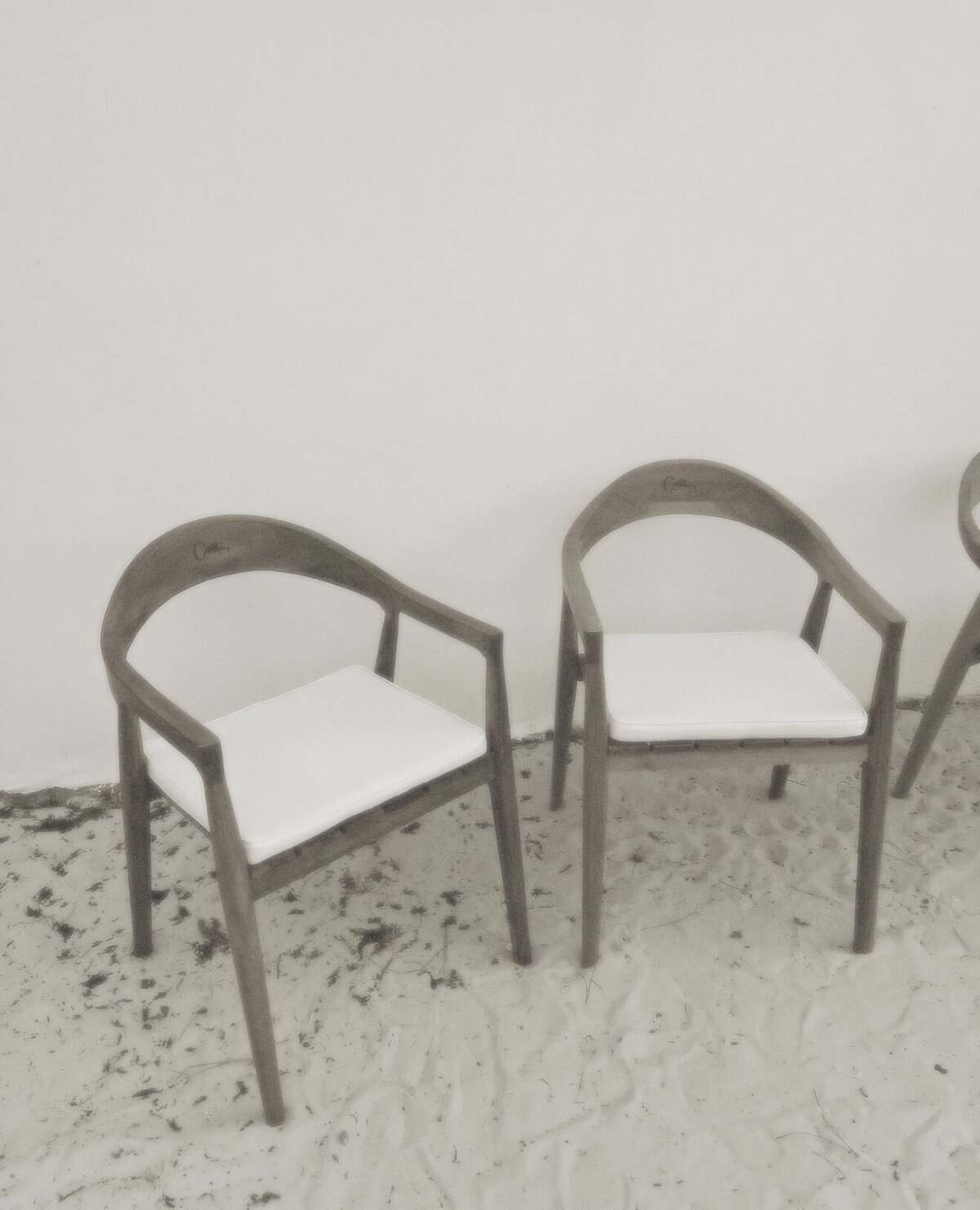Two wooden chairs with white cushions, positioned against a white wall. The chairs are minimalistic and modern with a curved backrest.