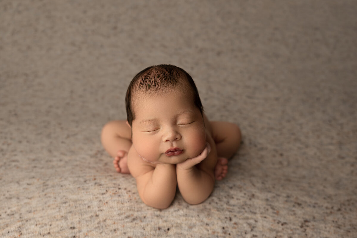 Fine art newborn baby photoshoot captured by the best New Jersey baby photographer Katie Marshall. Baby boy is sleeping bare and sitting in the froggy pose. His hands are propping his chin. Baby boy is laying on a marled beige stretch fabric.