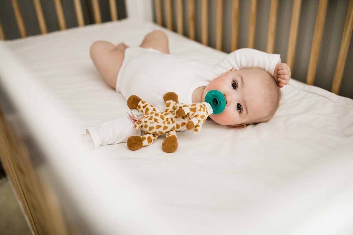 Infant with a pacifier lying in a crib, holding a giraffe toy, captured beautifully by a family photographer Pittsburgh.