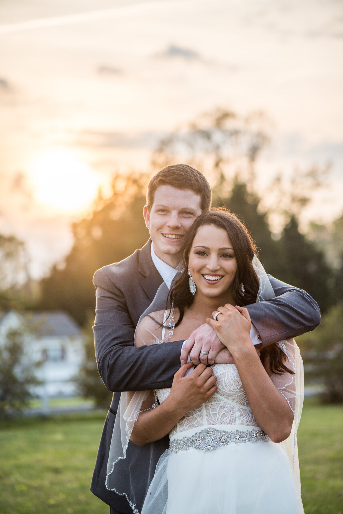 A person standing behind their partner with their arms around them as they both smile on their wedding day 