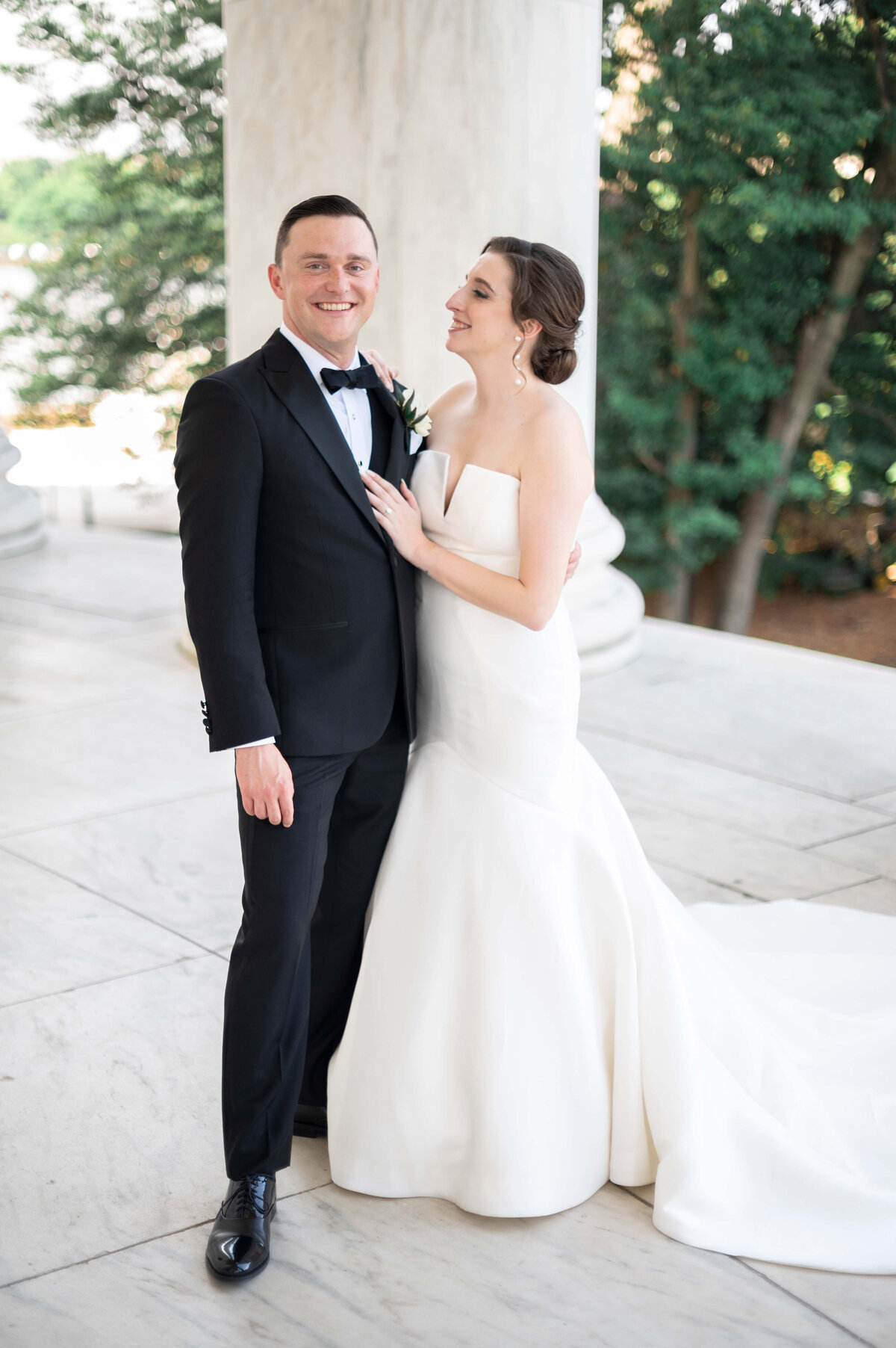 A bride in a strapless white gown and a groom in a black tuxedo stand together, smiling, on a marble terrace with trees in the background.