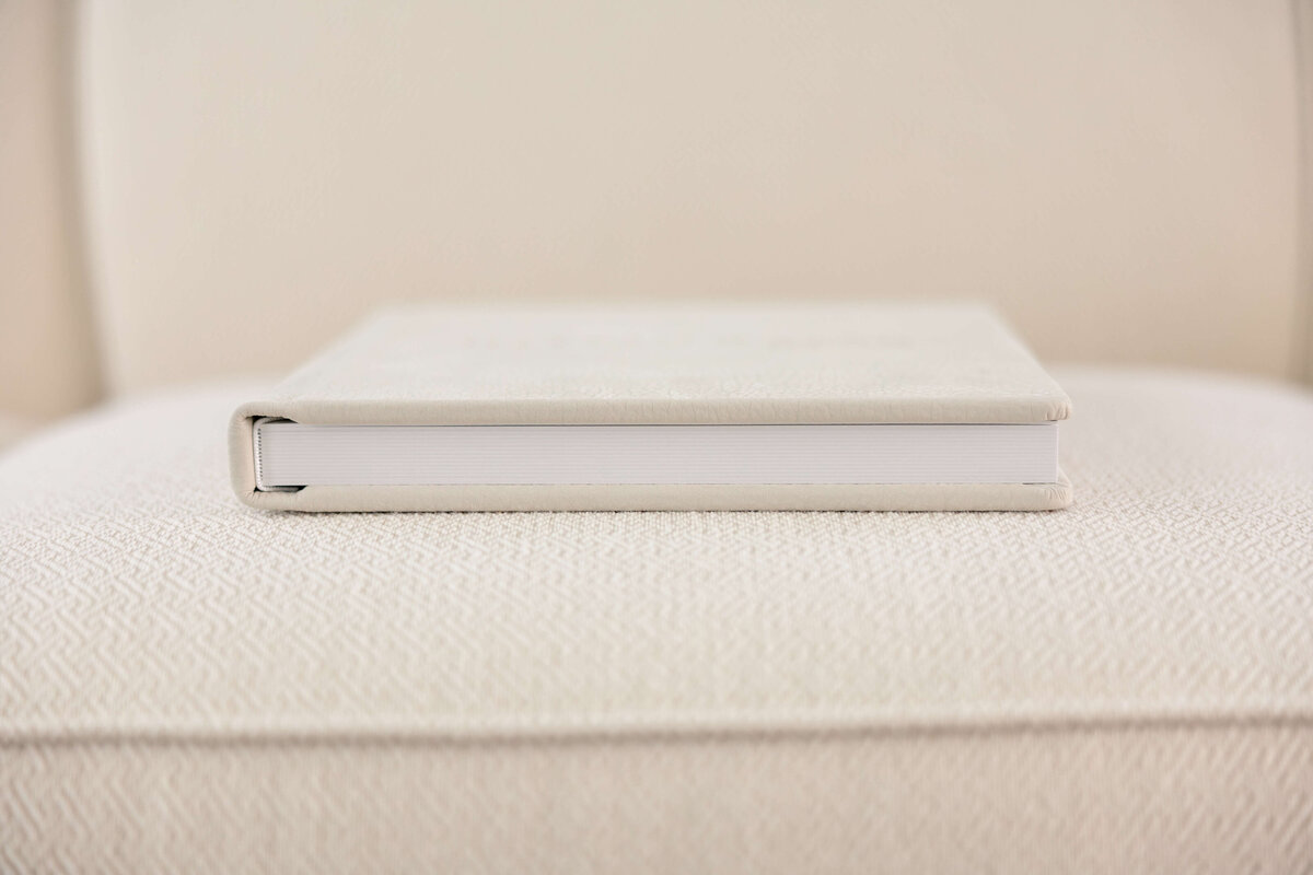 A closed, light-colored book rests horizontally on a matching textured surface, blending with the soft, neutral background. The books spine and white pages are visible, emphasizing a clean, minimalist aesthetic.