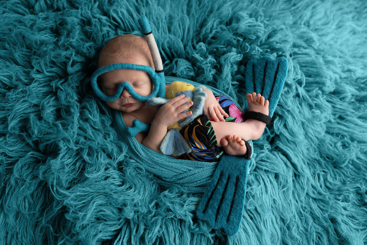 Adorable newborn boy posed in tropical pants with felt snorkel and flippers on a teal backdrop.