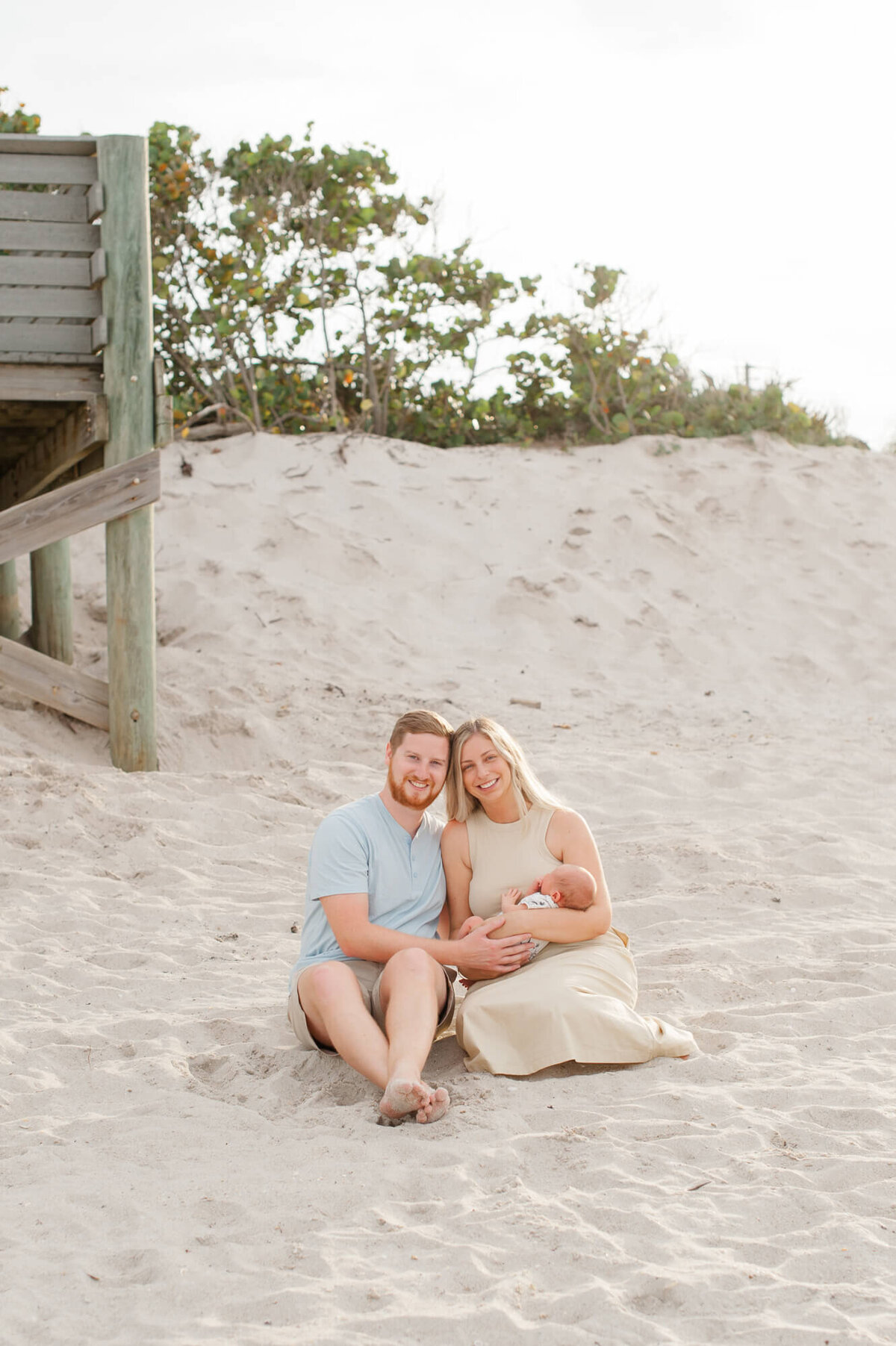 Parents smile at the camera while holding their newborn son in the sand