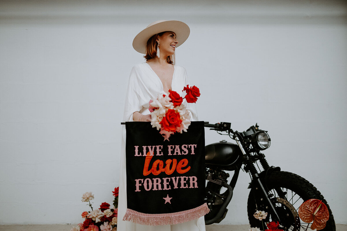 A boho bride stands in front of a bike holding a custom wedding banner.