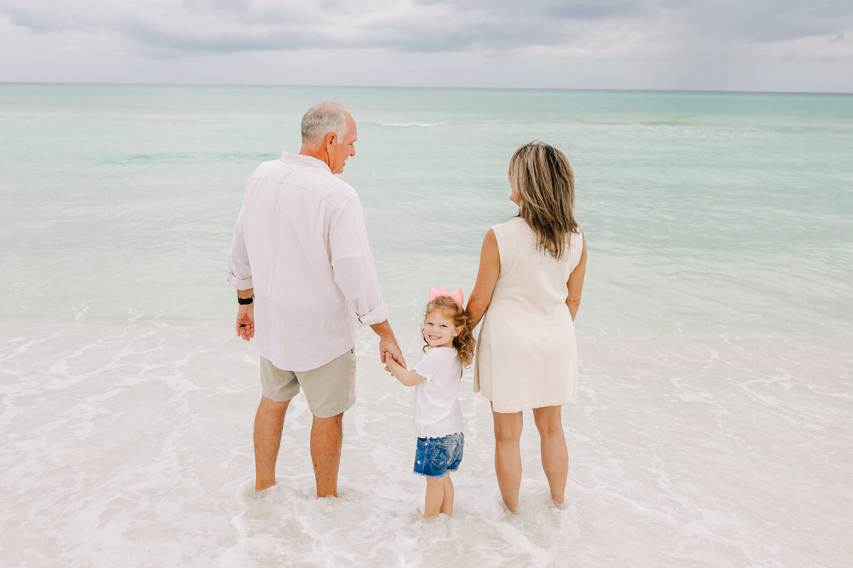A little girl with curly blonde hair and a pink bow holds hands with her grandparents as they stand in the gentle waves of Orange Beach, AL. She looks back with a bright smile, capturing a heartwarming multi-generational family moment by the ocean. The grandparents, dressed in light coastal attire, gaze out at the serene turquoise water. A perfect beach portrait celebrating family bonds and joyful memories by the shore.