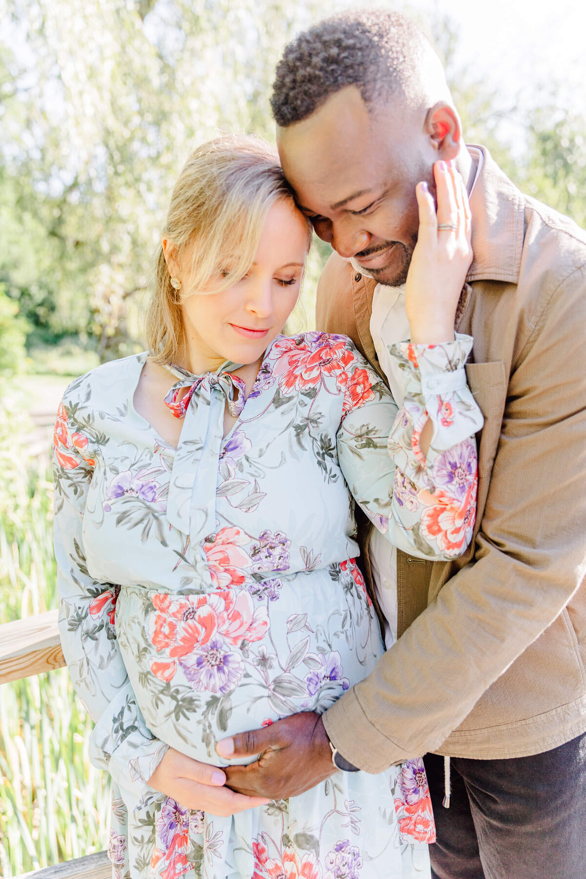 Pregnant woman in light floral dress and her husand gaze down at her bump while cradling it together
