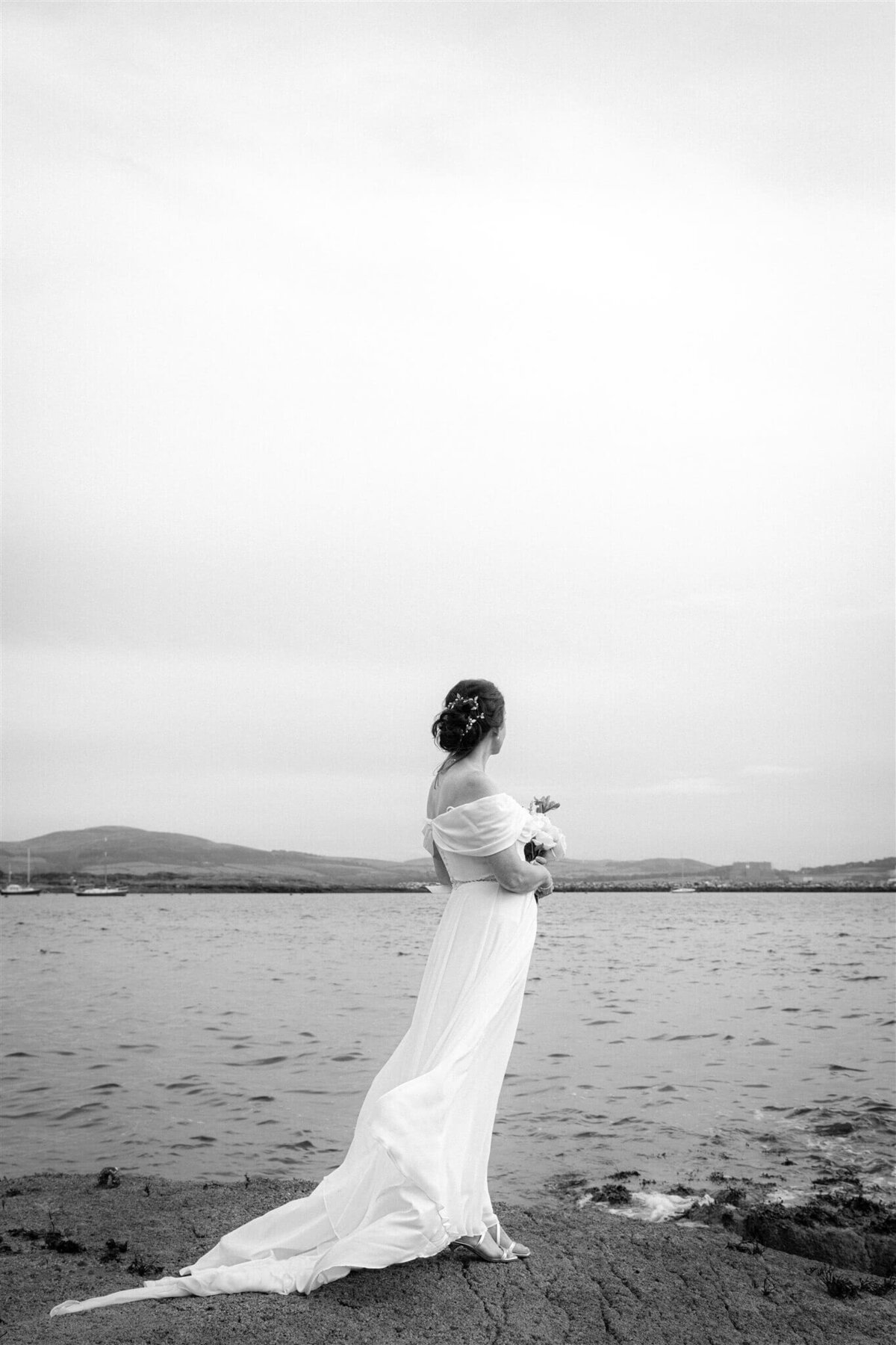 A bride holds a bouquet and looks out to sea. Hills on the distant landscape.