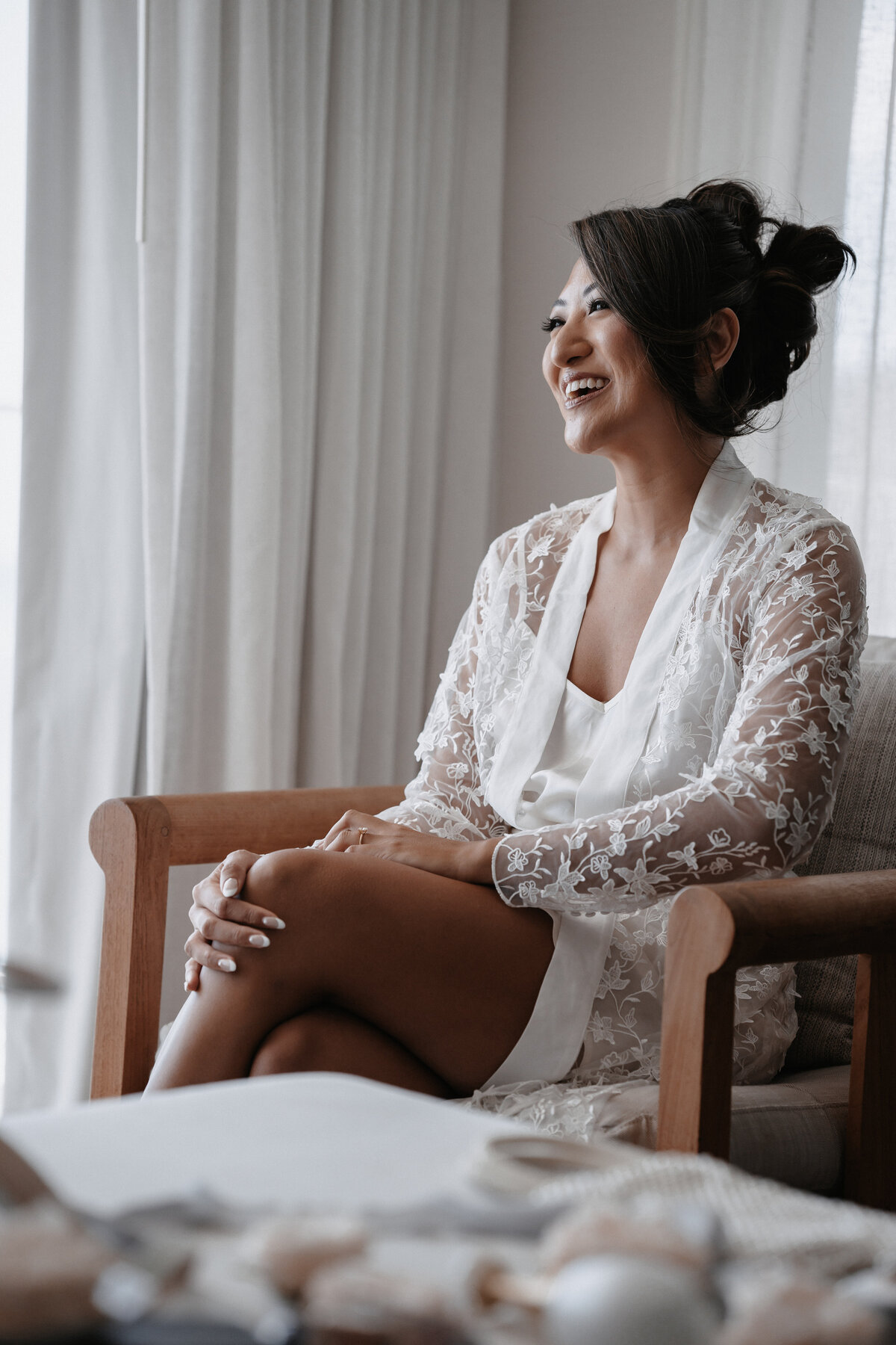 Bride seated in lace robe with legs crossed laughing