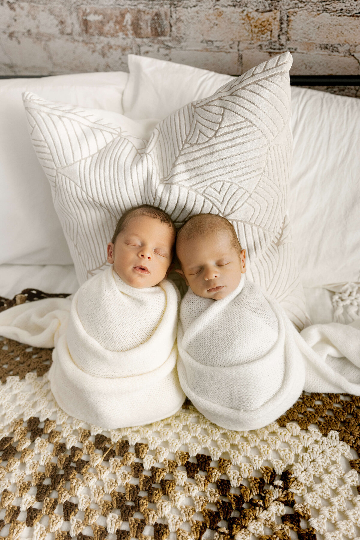 Two newborn babies, swaddled in white blankets, are sleeping closely together on a textured, multi-colored blanket. The background features soft pillows, creating a warm and serene atmosphere.