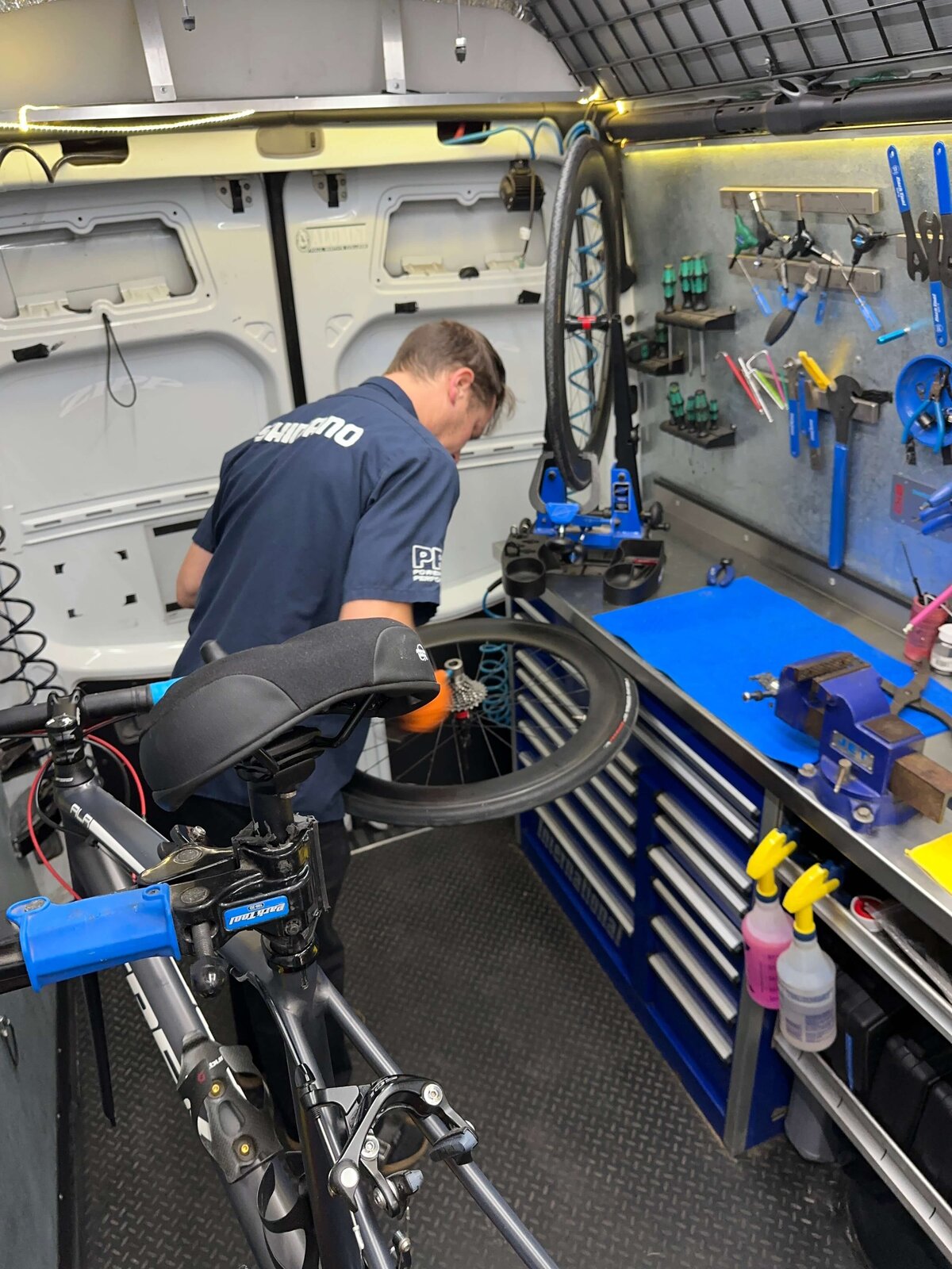 A bike mechanic works inside a well-organized mobile repair van, fixing a bicycle wheel. The vans interior has tools hanging neatly on the wall, a blue workbench, and multiple drawers. The mechanic wears a blue shirt and gloves