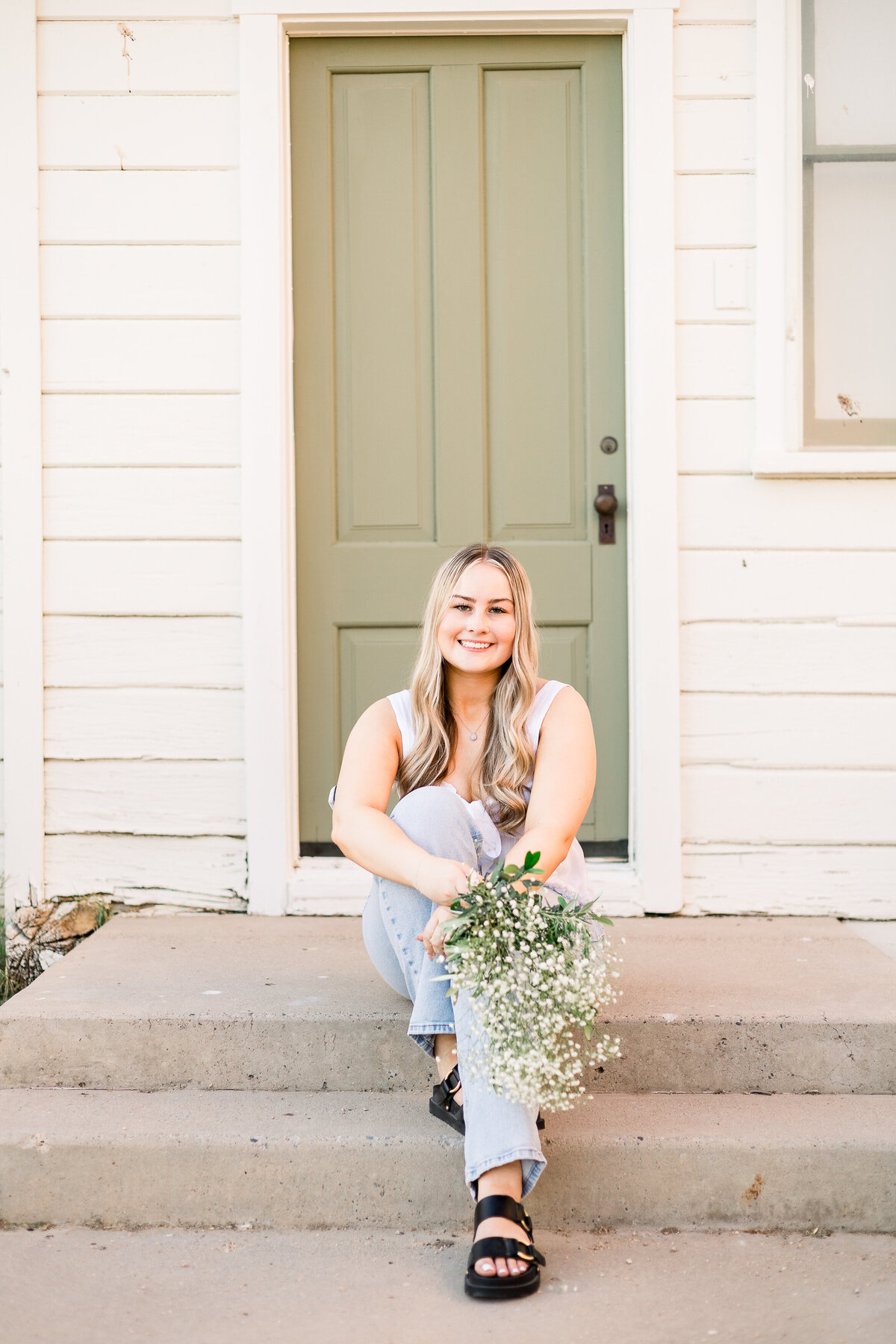 Senior sits on doorstep in front of green door on historic building at Sahuaro Ranch Park in Glendale, AZ, holding a bouquet of baby's breath.