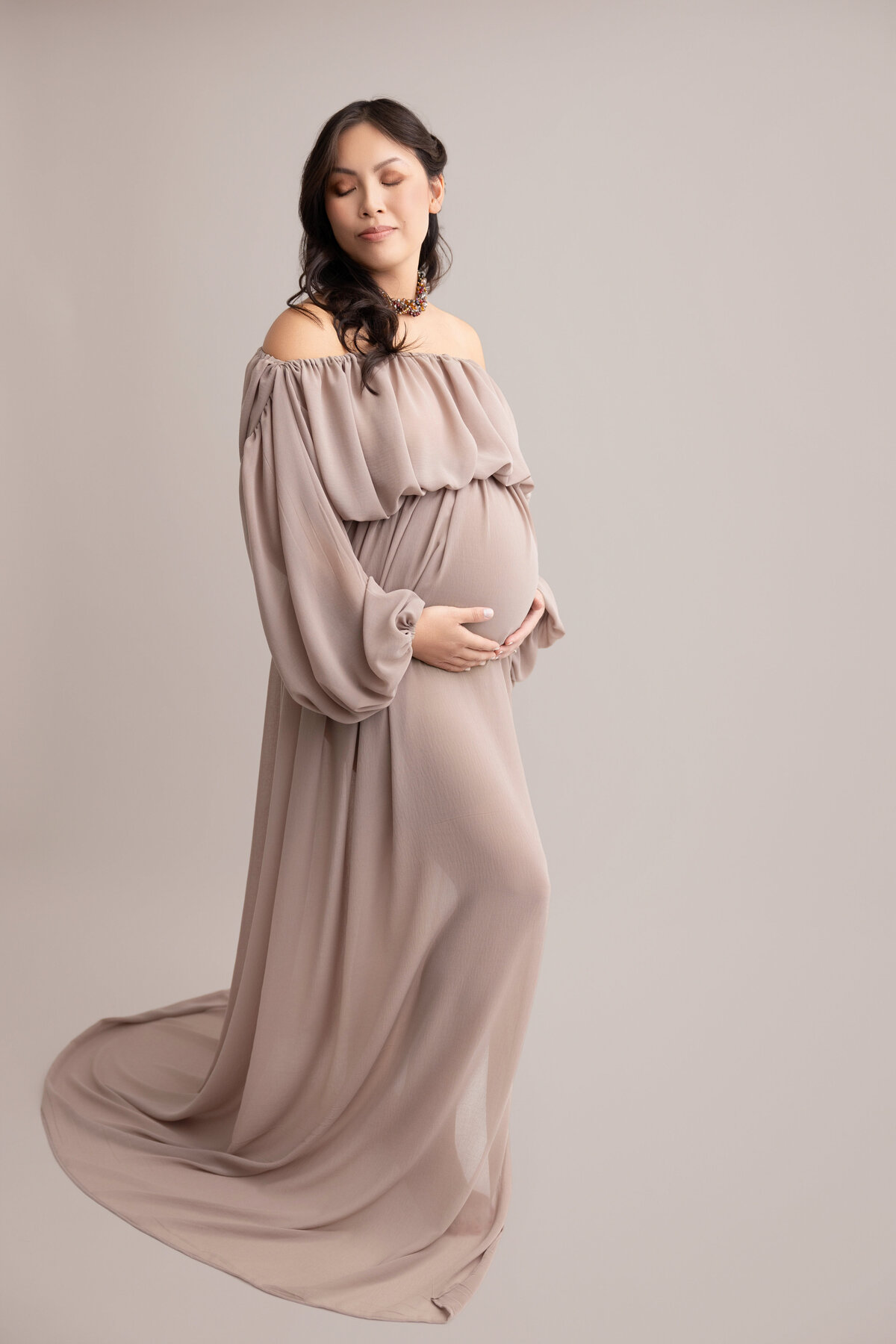 Maternity image of pregnant mom wearing a brown airy dress and posed with hands under the belly against a light brown backdrop.
