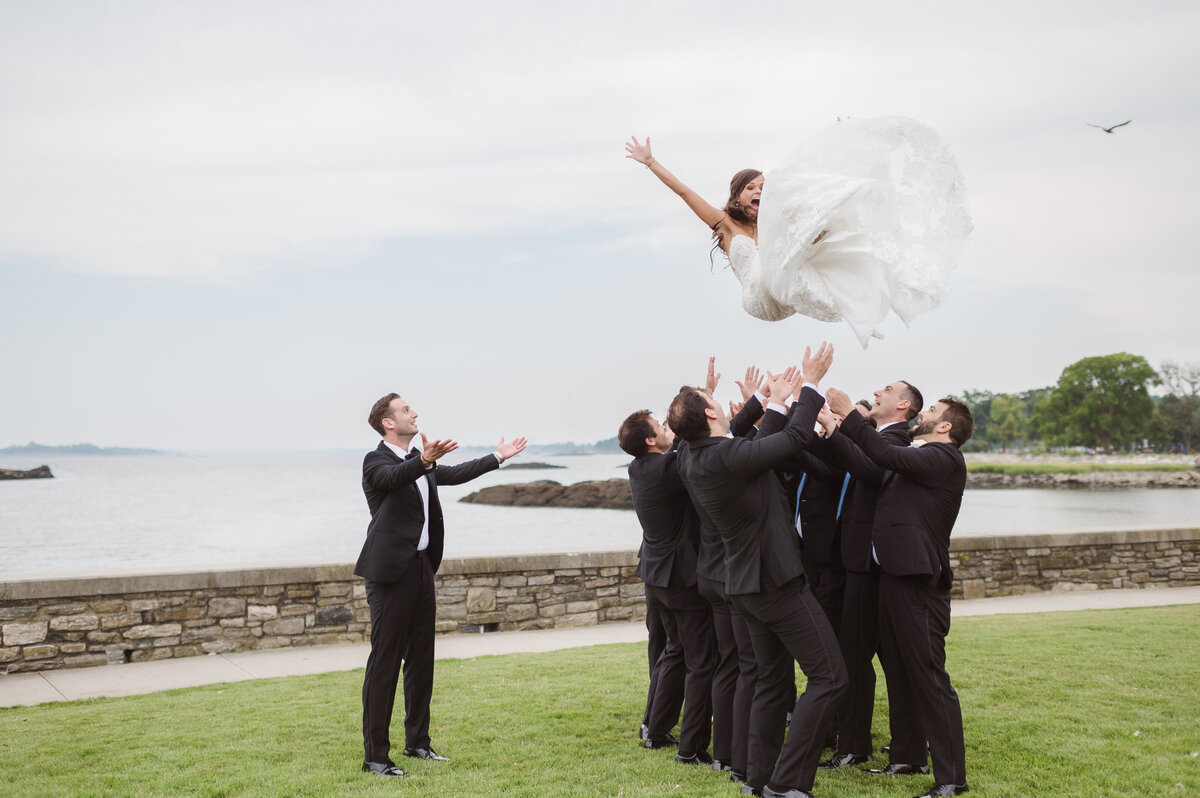 A funny wedding party group shot of the groomsmen tossing the bride in the air like a cheerleader