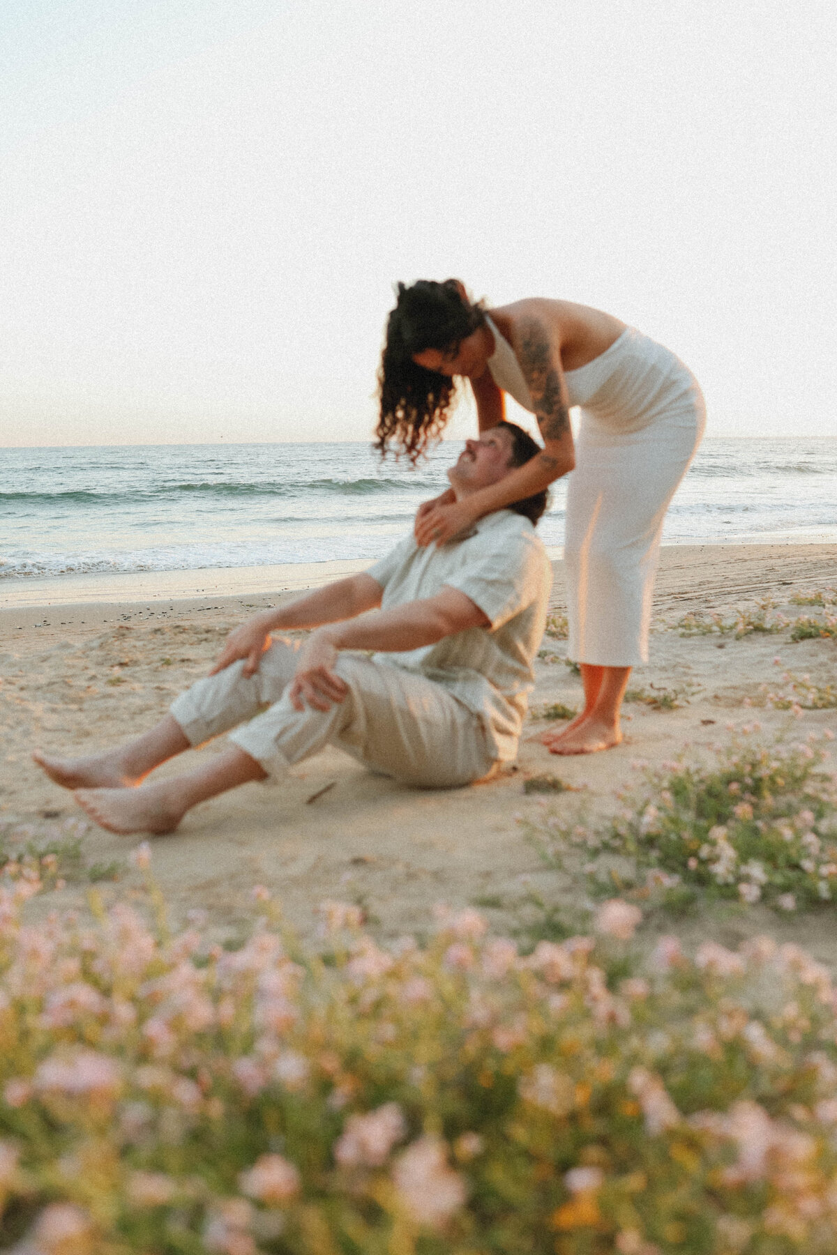 man sitting on the beach while girlfriend leans over him and kisses him with the ocean in the back round