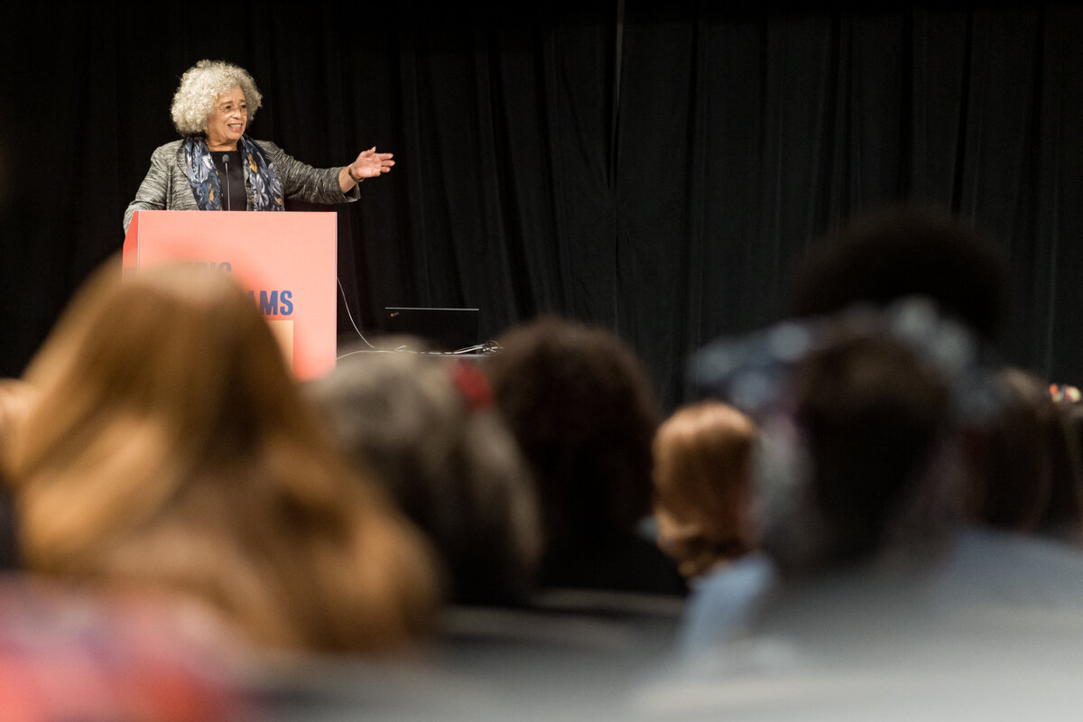 a woman with gray hair stands in front of a podium, holding out an arm