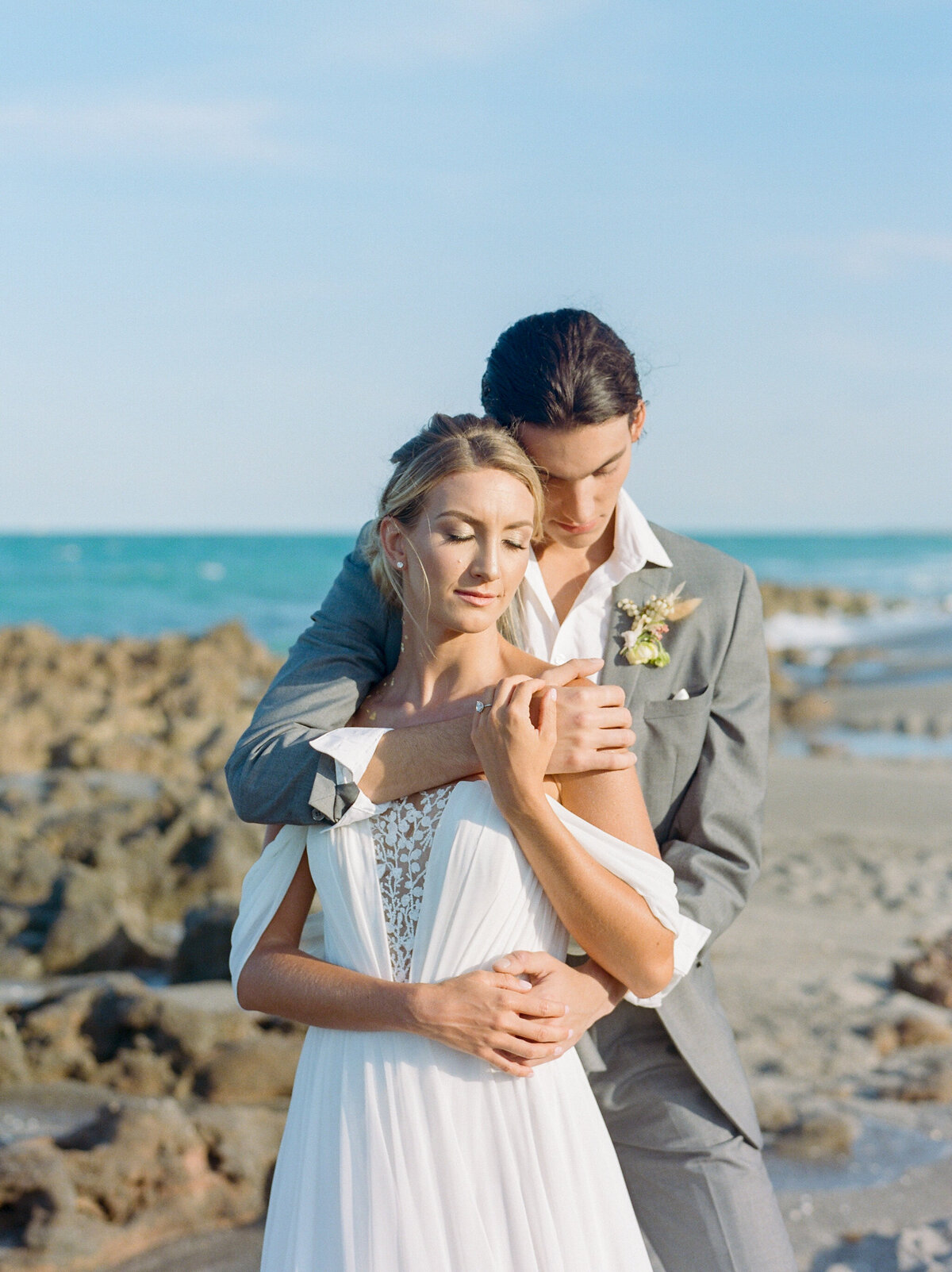 Groom holding bride from behind at beach.