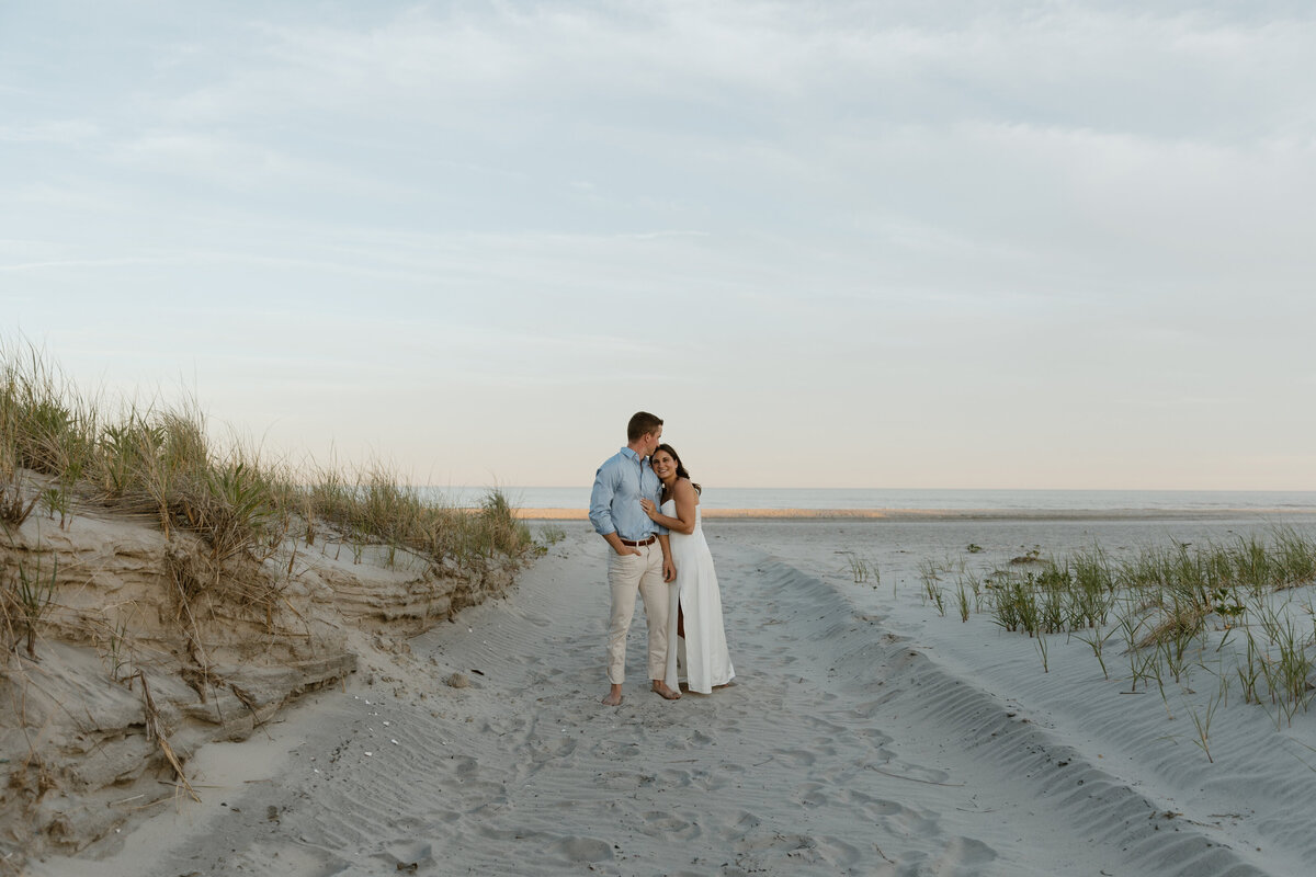 A couple stands on a sandy beach, embracing among gentle dunes and grass, with the serene ocean and sky in the background.
