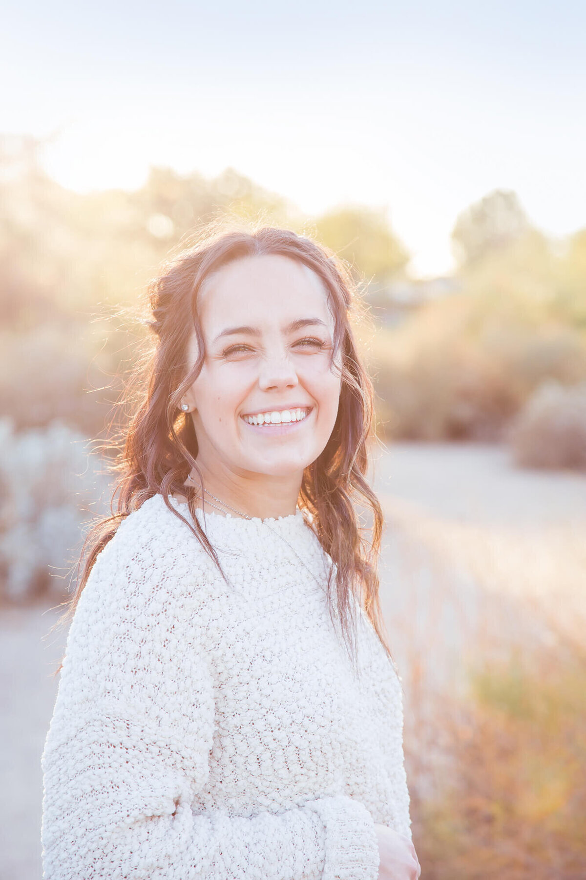 joyful brown haired young woman in white sweater on desert path with hazy sunset