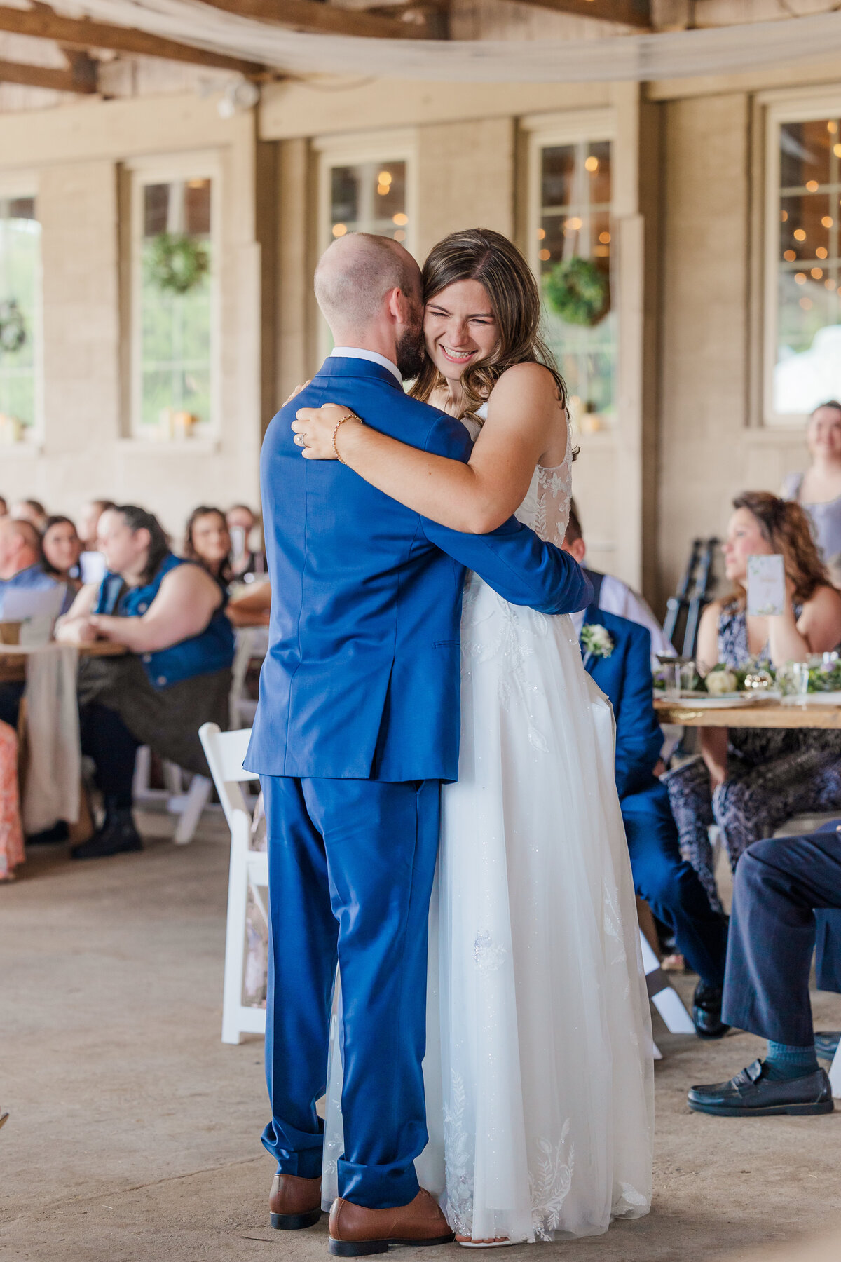 bride and groom sharing first dance wearing blue suit and white dress with family watching in background