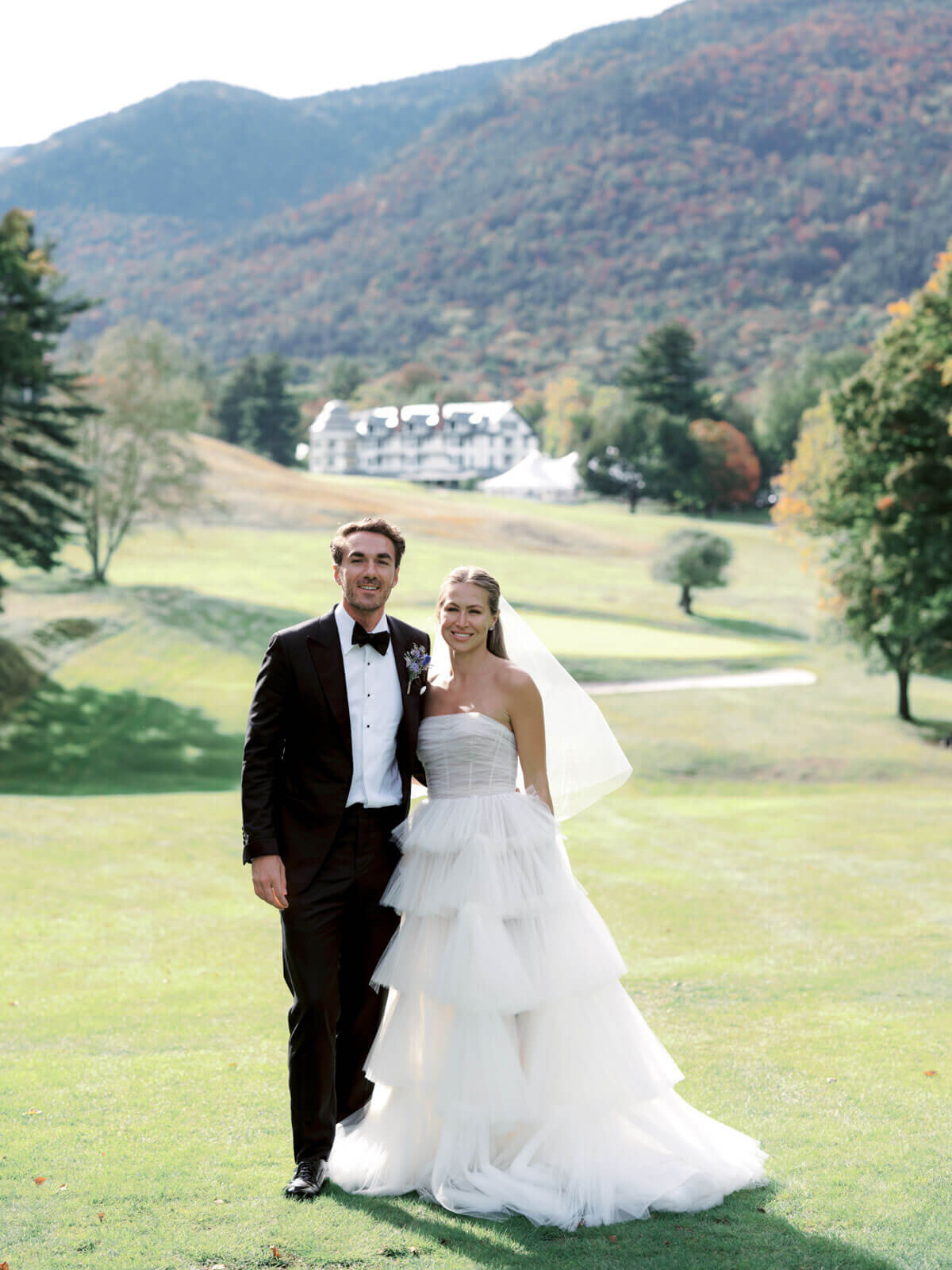 The bride and groom are standing close, smiling at the camera, at The Ausable Club's golf course, NY. Image by Jenny Fu Studio.