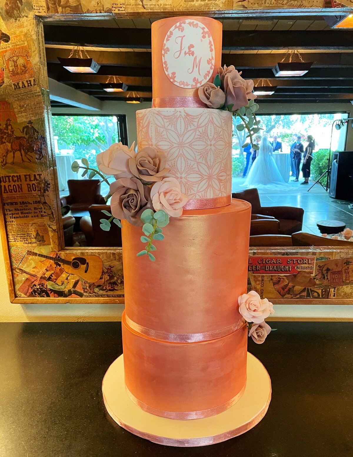Tall 4 tier rose gold wedding cake. Top tier with sugar roses and decorative monogram plaque; 2 nd tier is white with rose gold abstract leaf stencil; 3rd and bottom tiers are rose gold with sugar roses , eucalyptus accents