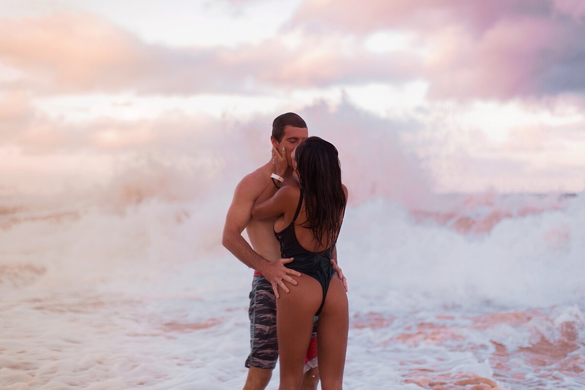Engagement portraits in Maui featuring a man holding his wife who wears a black one piece swimsuit