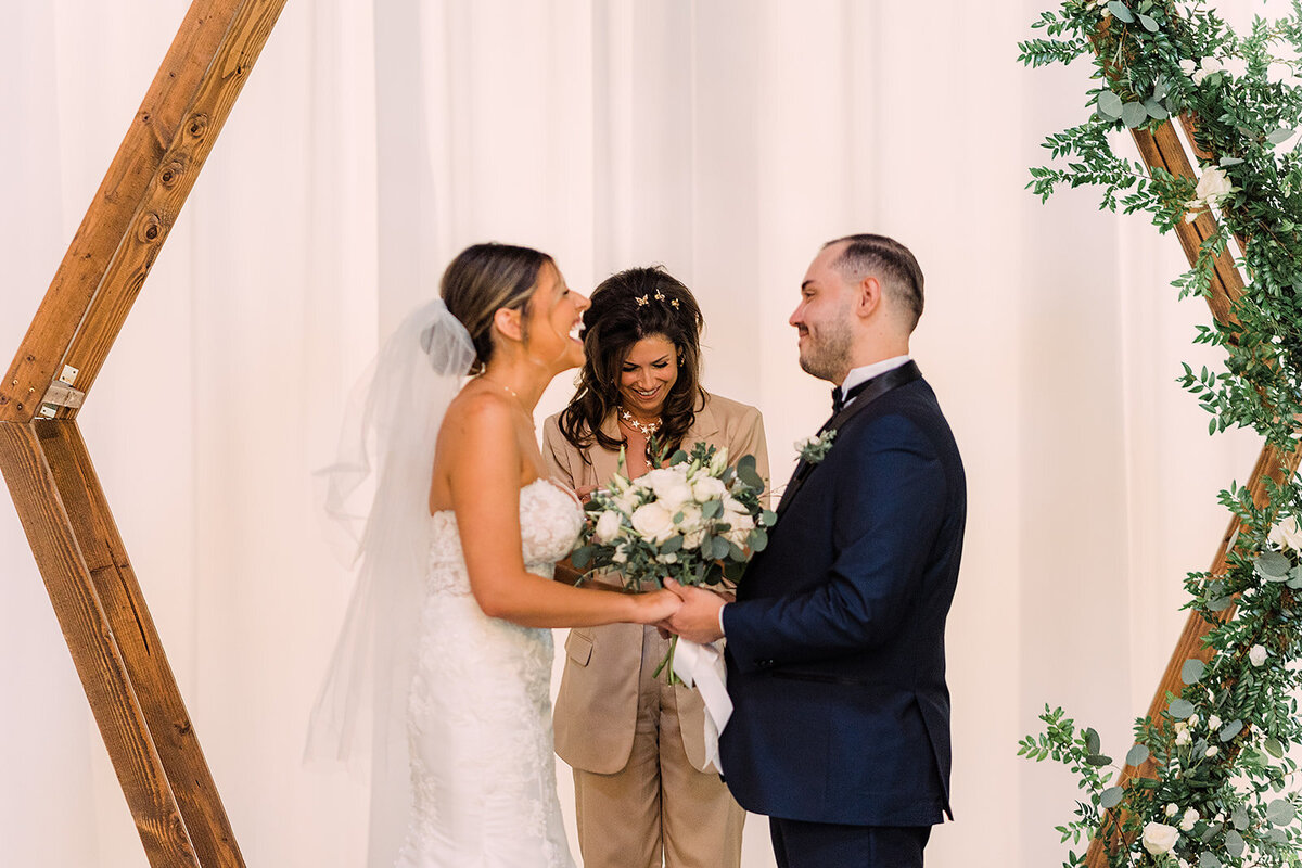 The bride and groom stand under a wooden arch decorated with greenery and white flowers, smiling at each other while the officiant behind them shares in their joyful moment.