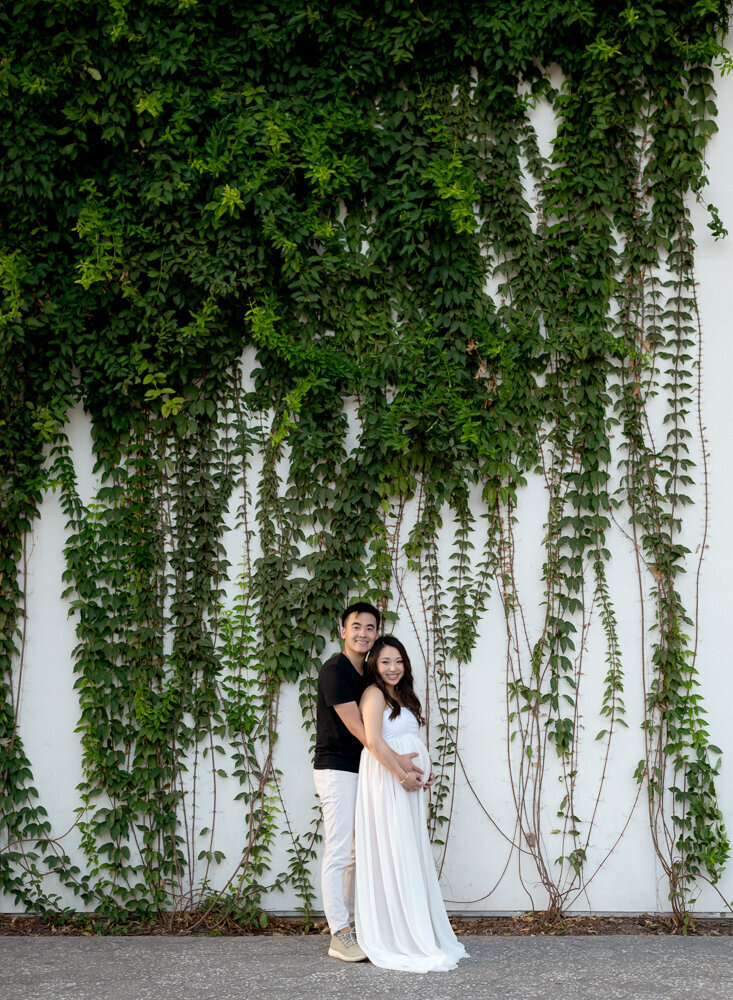 Maternity couple posing in front of a white wall covered in lush green vines during an outdoor session in Austin, Texas.