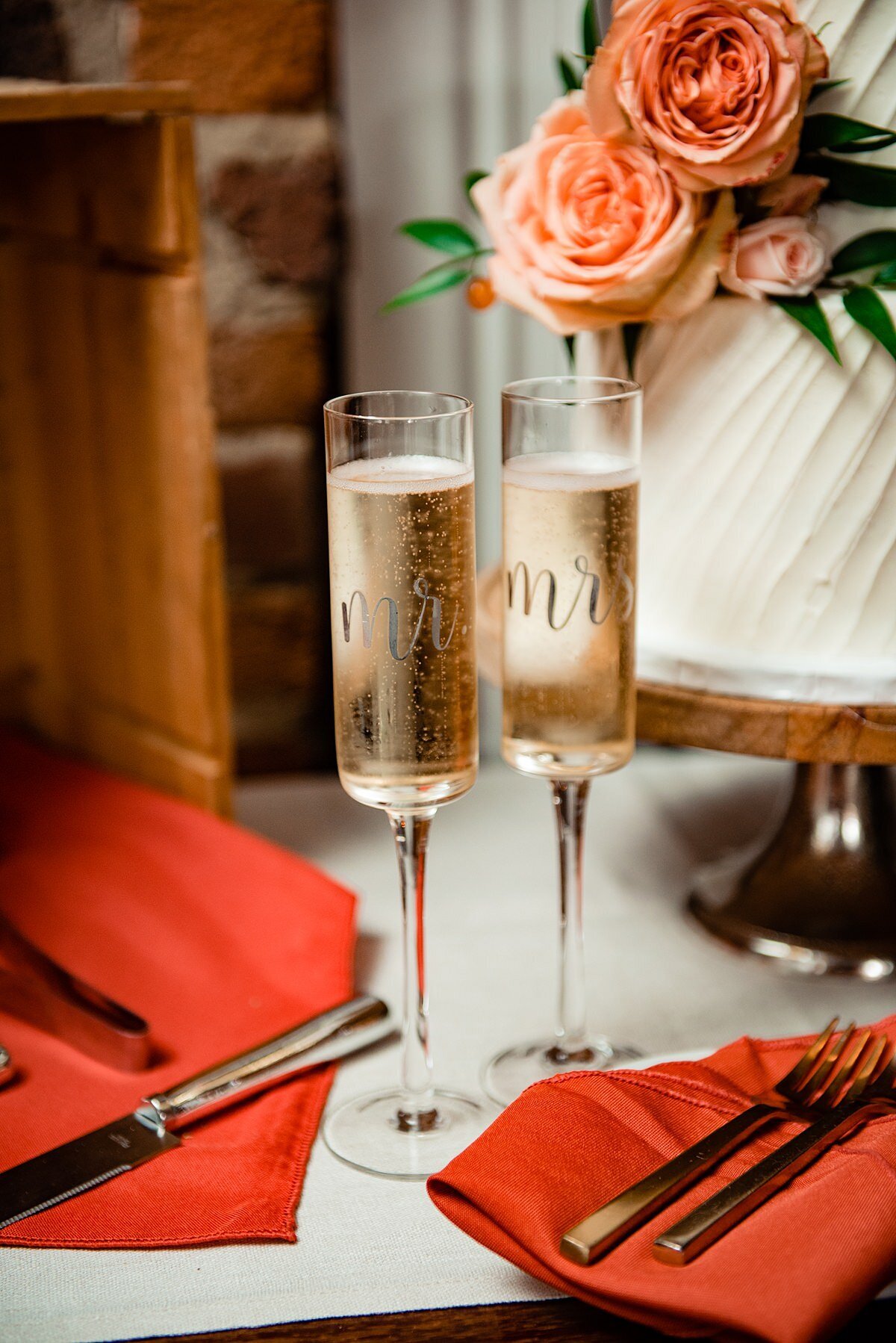 Mr. and Mrs. champagne flutes filled with champagne sit between two burnt orange napkins with gold flatware. Behind the glasses is the two tier white wedding cake with striped frosting and blush roses.