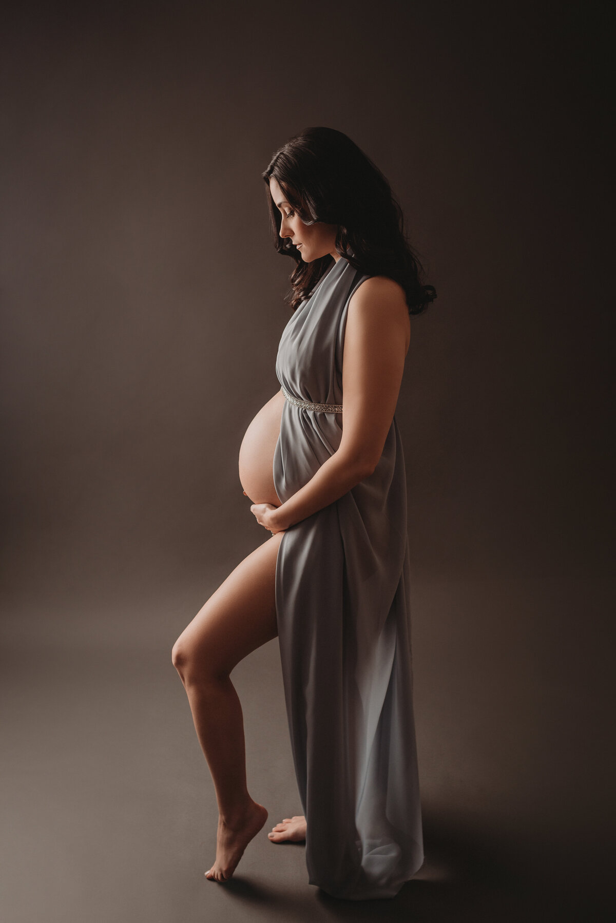 Moody pregnancy portrait with 35 weeks pregnant woman in jade blue chiffon fabric standing to side holding and looking at baby bump
