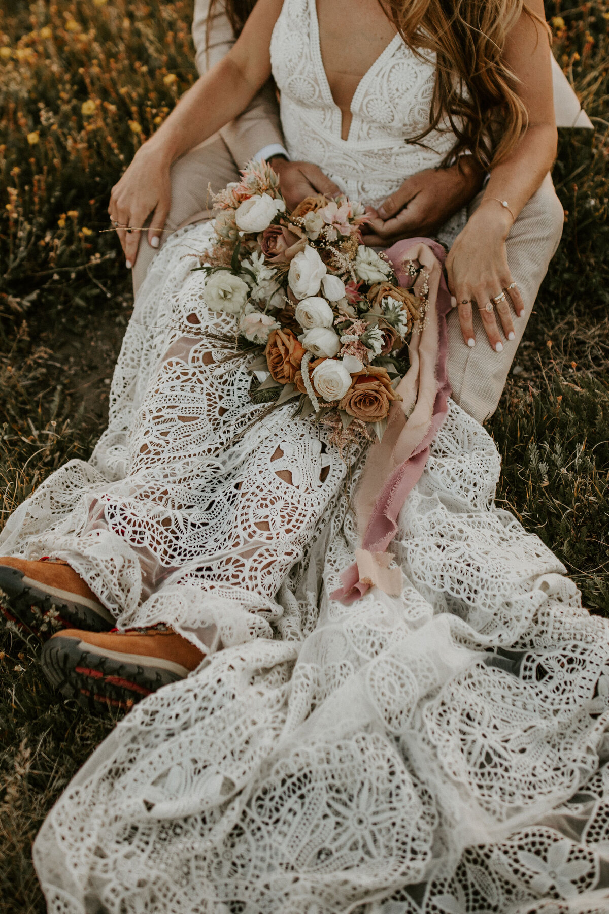 bride is hugged by groom while wearing a white wedding gown and hiking boots, holding a bouquet sitting on grass